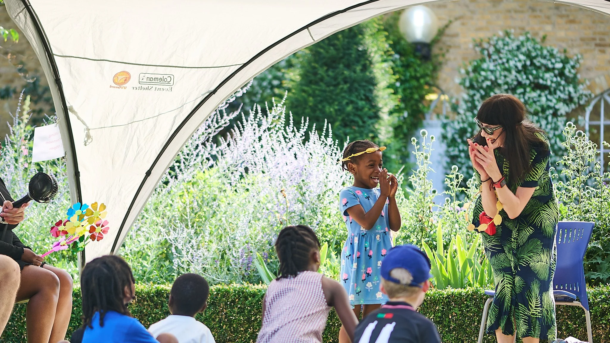 Children and an adult woman engaging in a storytelling activity outdoors under a canopy, with plants and flowers around.