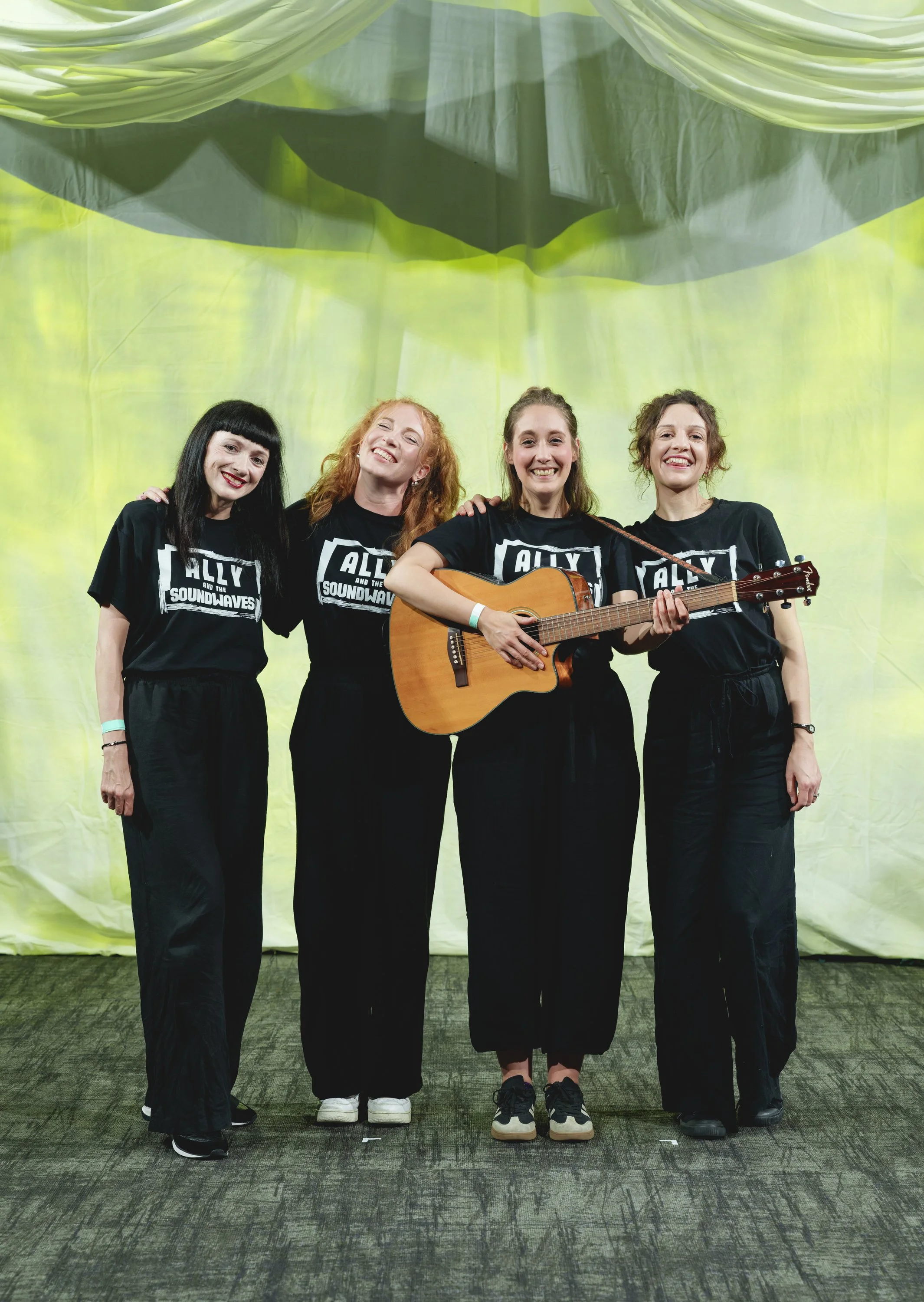 Four women standing together on a stage, smiling, with a green abstract backdrop; all wearing black T-shirts with white text that reads 'ALLY AND THE SOUNDWAVES' and black pants. They are performers and facilitators for a sensory experience.
