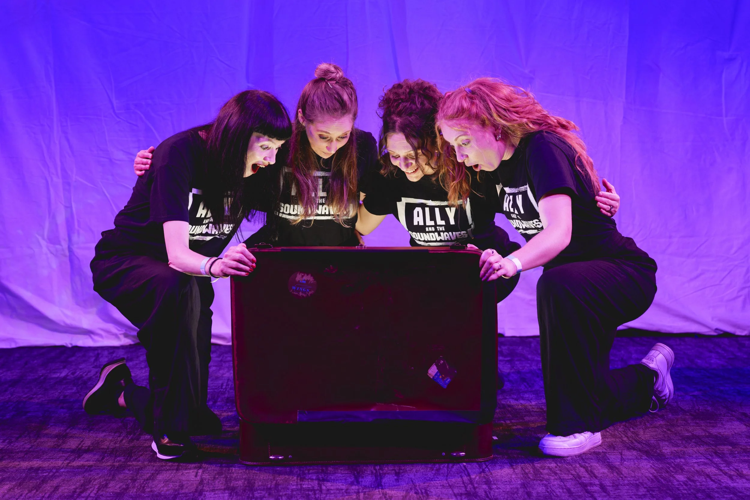 Five women wearing black T-shirts with 'Ally and the Soundwaves' on them, crouching around a glowing, brown suitcase, looking inside with excitement, against a purple background. They are performing a sensory show.