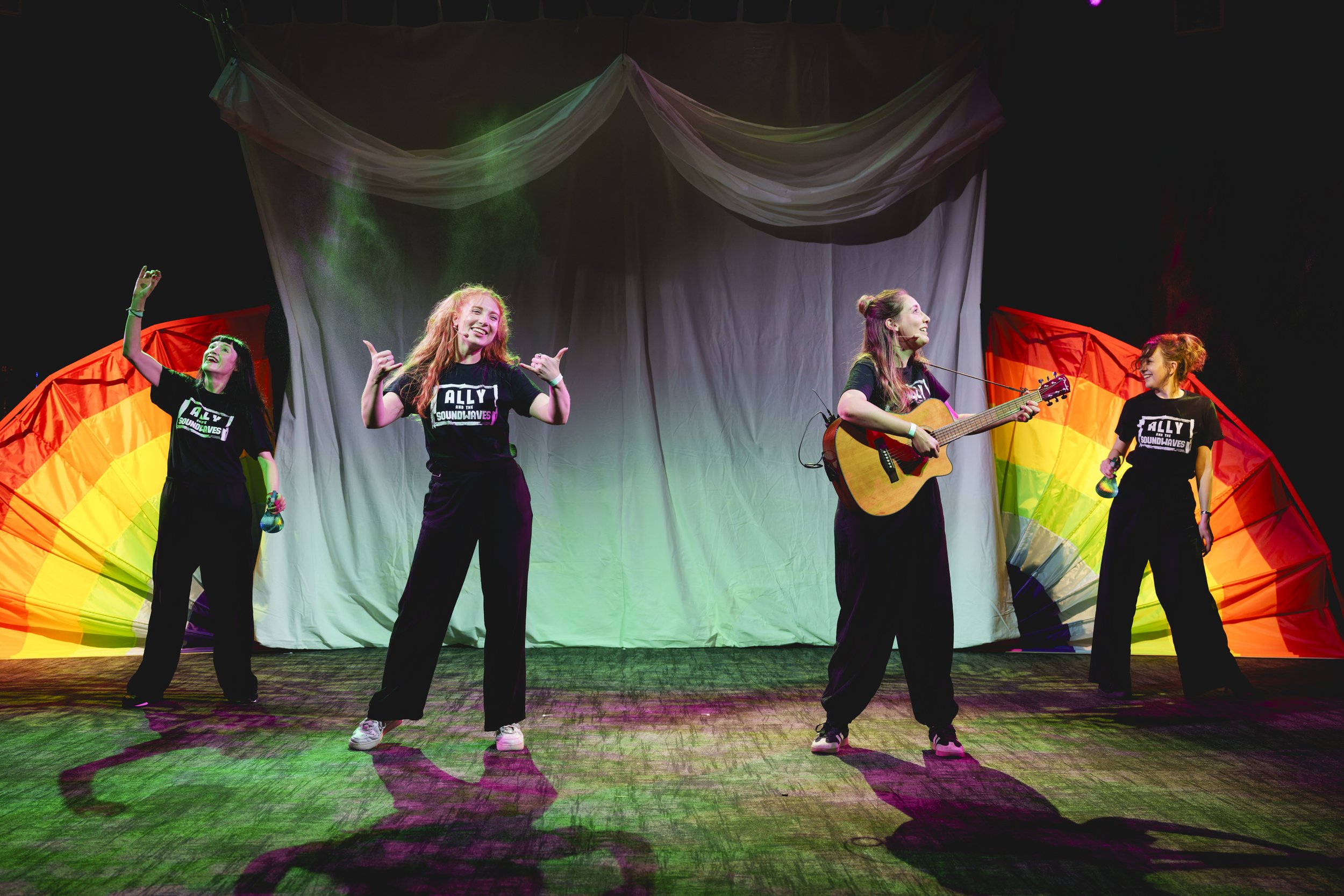 Four young female workshop facilitators performing onstage at the Royal Albert Hall,  for autistic and/or learning-disabled children, with a rainbow and green backdrop. One woman is playing guitar, others are singing or dancing, all smiling.