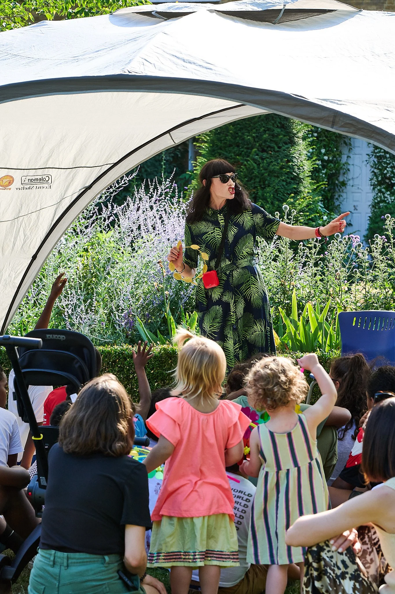 Woman in sunglasses and tropical dress telling a fun story to a group of children outdoors with a garden background