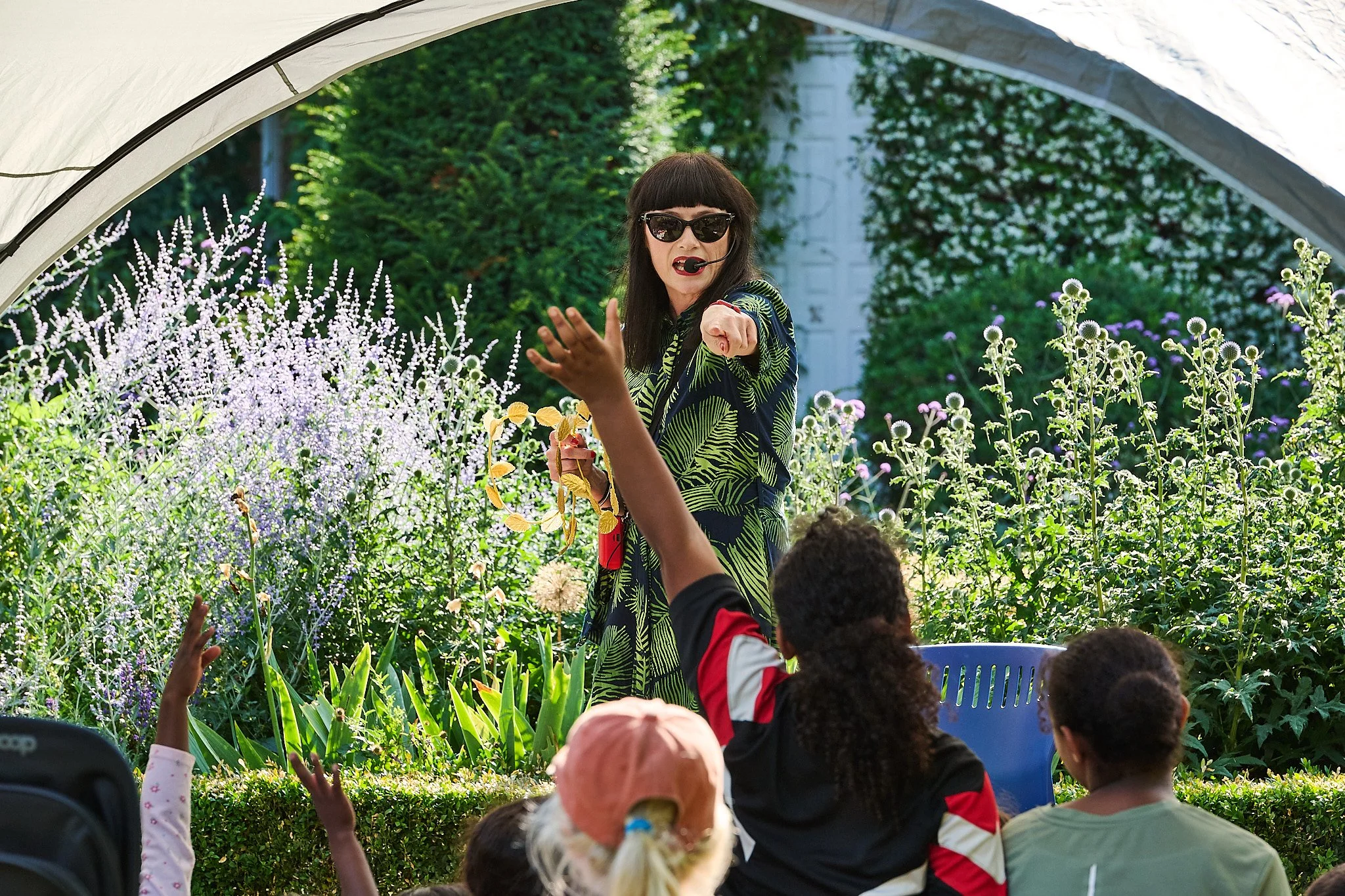 A woman with dark hair, sunglasses, and a colourful dress is story telling under a canopy outside. She is using  a microphone. There are flowers and lots of green grass.  Children in the audience raise their hands.