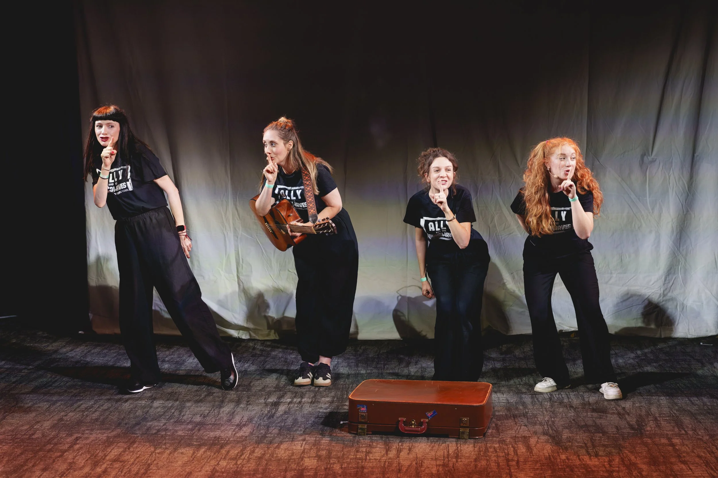 Four young women performing and facilitating on stage with a dark background. They are dressed in casual black t-shirts and pants, with one holding a guitar. All are making a gesture with one finger to their lips, suggesting a secret or quiet theme.