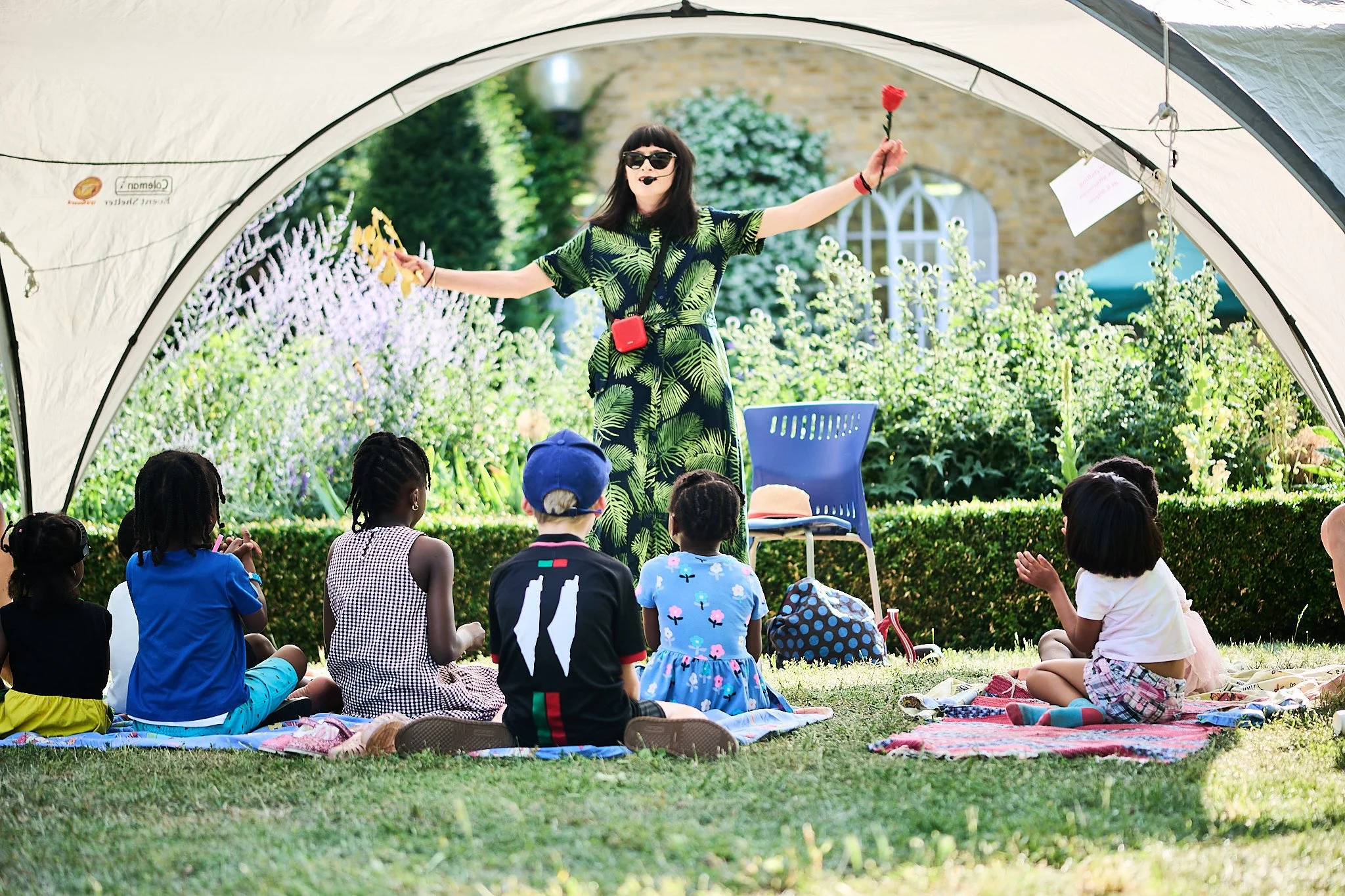 A woman storytelling, standing in a dramatic way,  holding a red flower while wearing sunglasses, with children sitting on blankets watching outdoors under a tent. It is a sunny day and an exciting story.