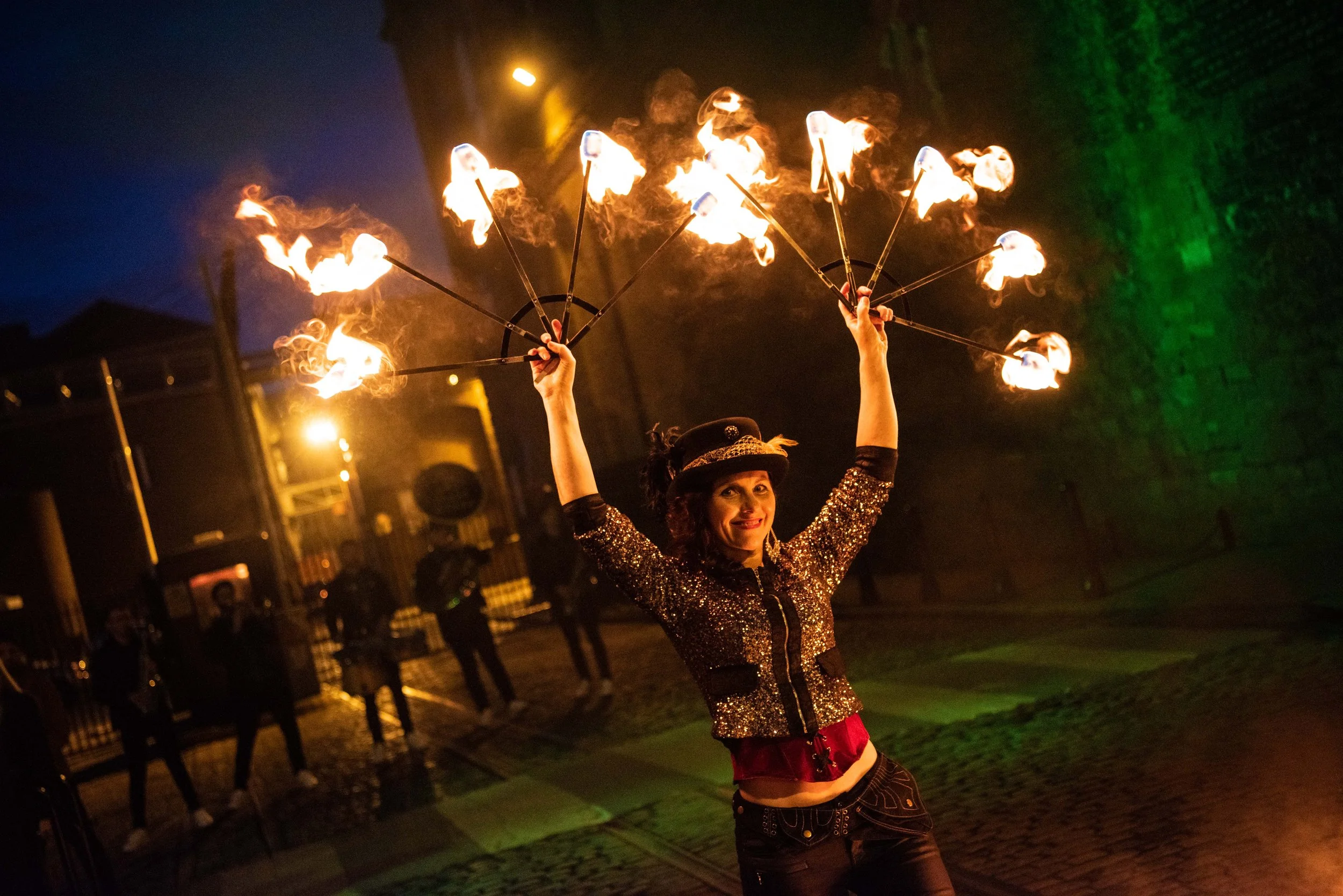 A four-piece folk band performs with a bodhrán, fiddle, banjo, and guitar against a vibrant, multi-colored geometric mural. The scene captures a lively, warm-lit atmosphere with the blurred silhouettes of a crowded audience in the foreground.