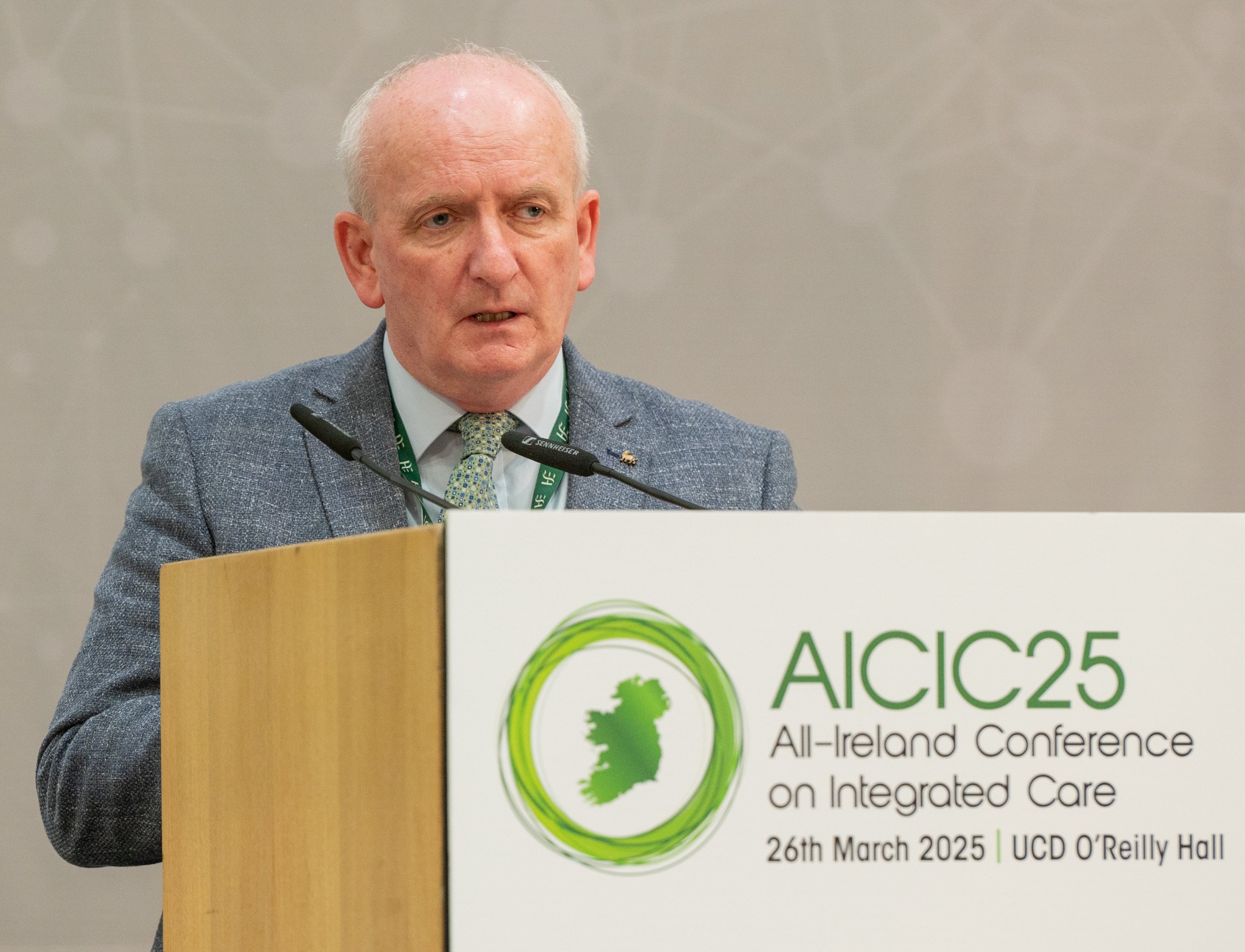 A man in a blue textured blazer and green patterned tie speaks from a wooden lectern featuring the "AICIC25" conference logo. He is positioned in front of a neutral, patterned backdrop during the All-Ireland Conference on Integrated Care.