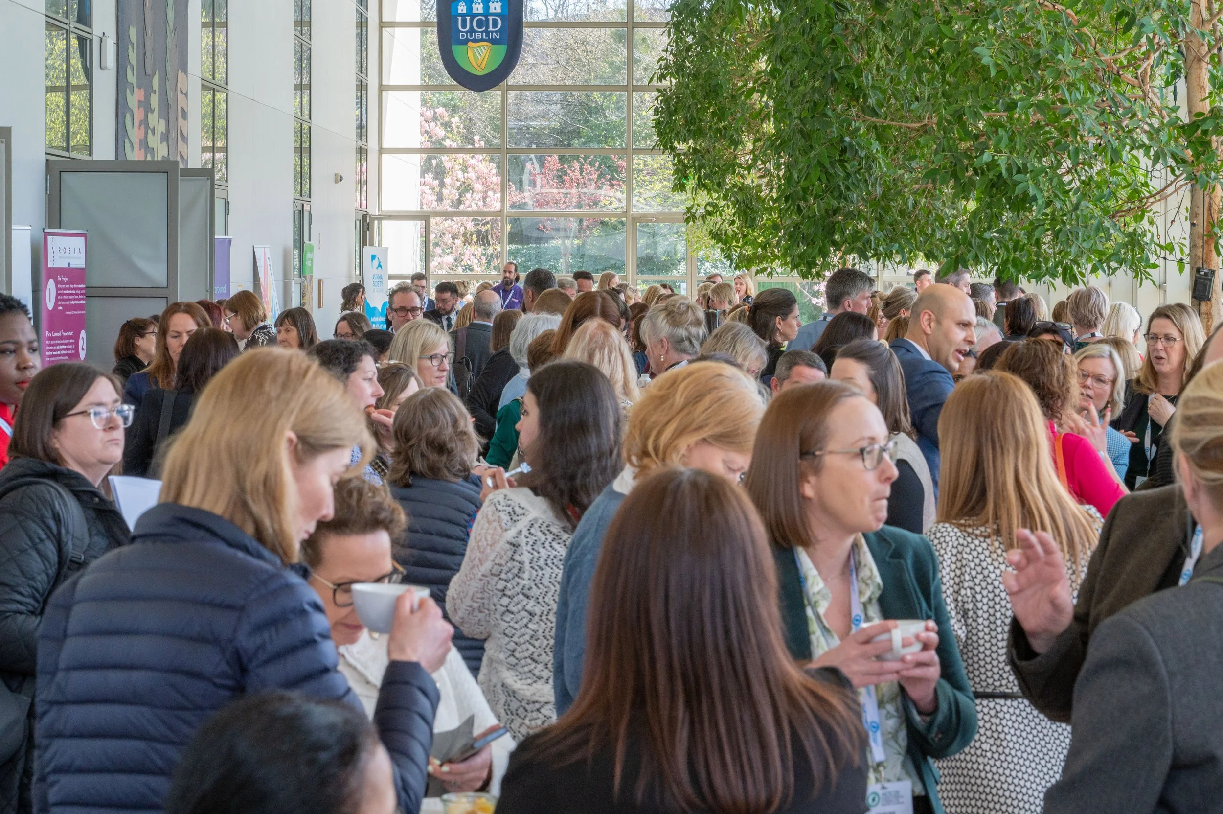 A large group of conference attendees gathers in a bright, modern atrium at UCD Dublin for a networking break with coffee. Large windows and indoor trees create a natural, airy environment as participants engage in conversation.