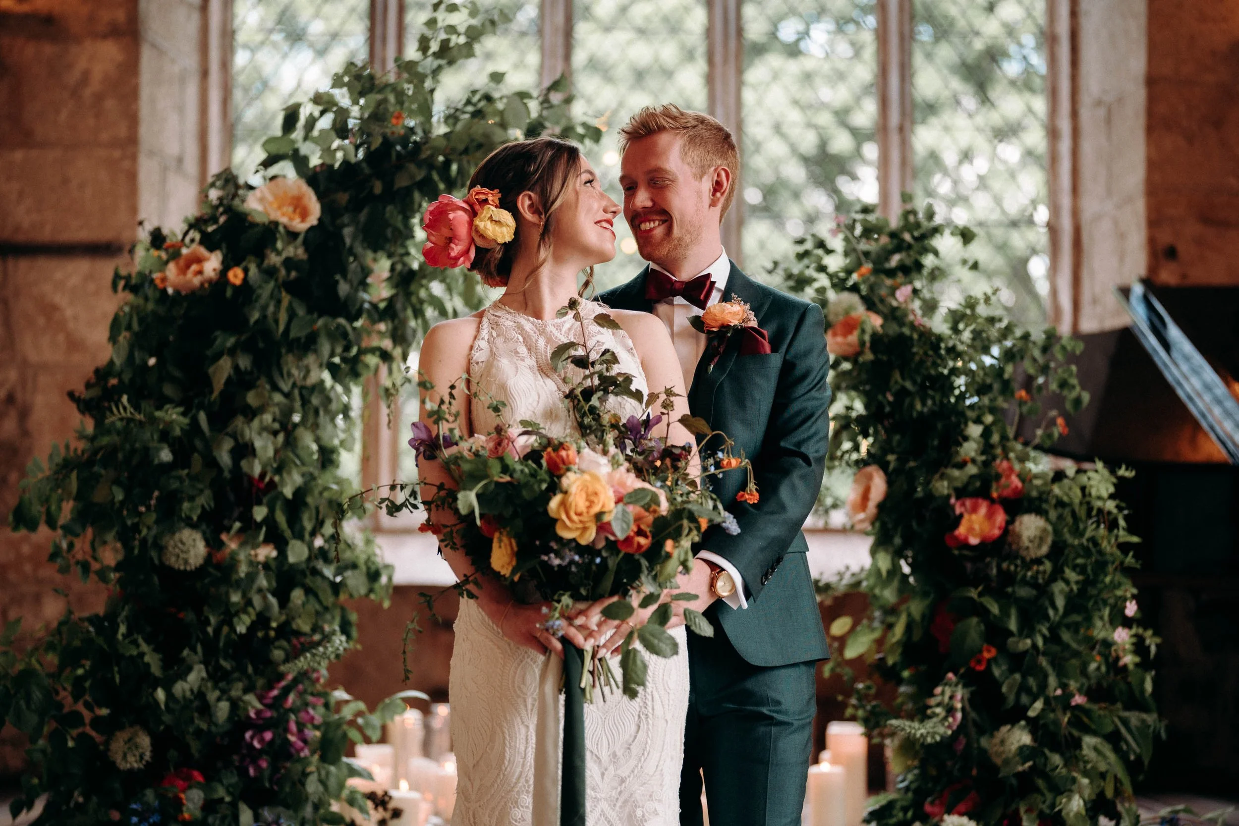Bride and groom standing inside The Priest’s House surrounded by lush floral arrangements, smiling at each other during their wedding portraits.