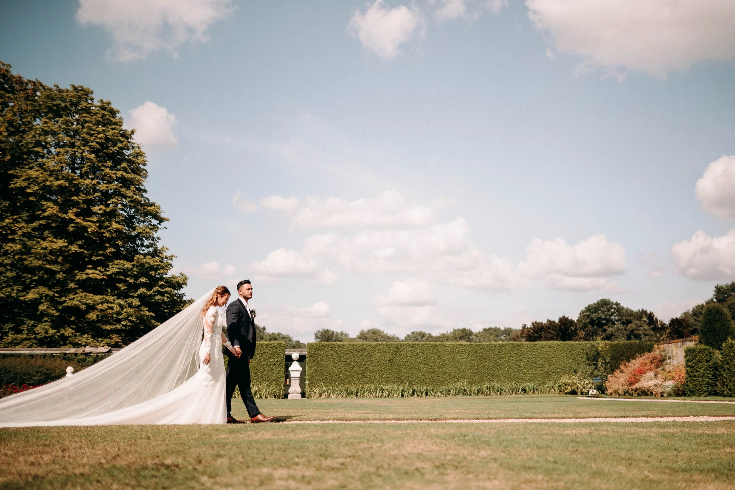 Bride and groom walking hand in hand across the gardens under a bright sky at Kasteel Heeswijk