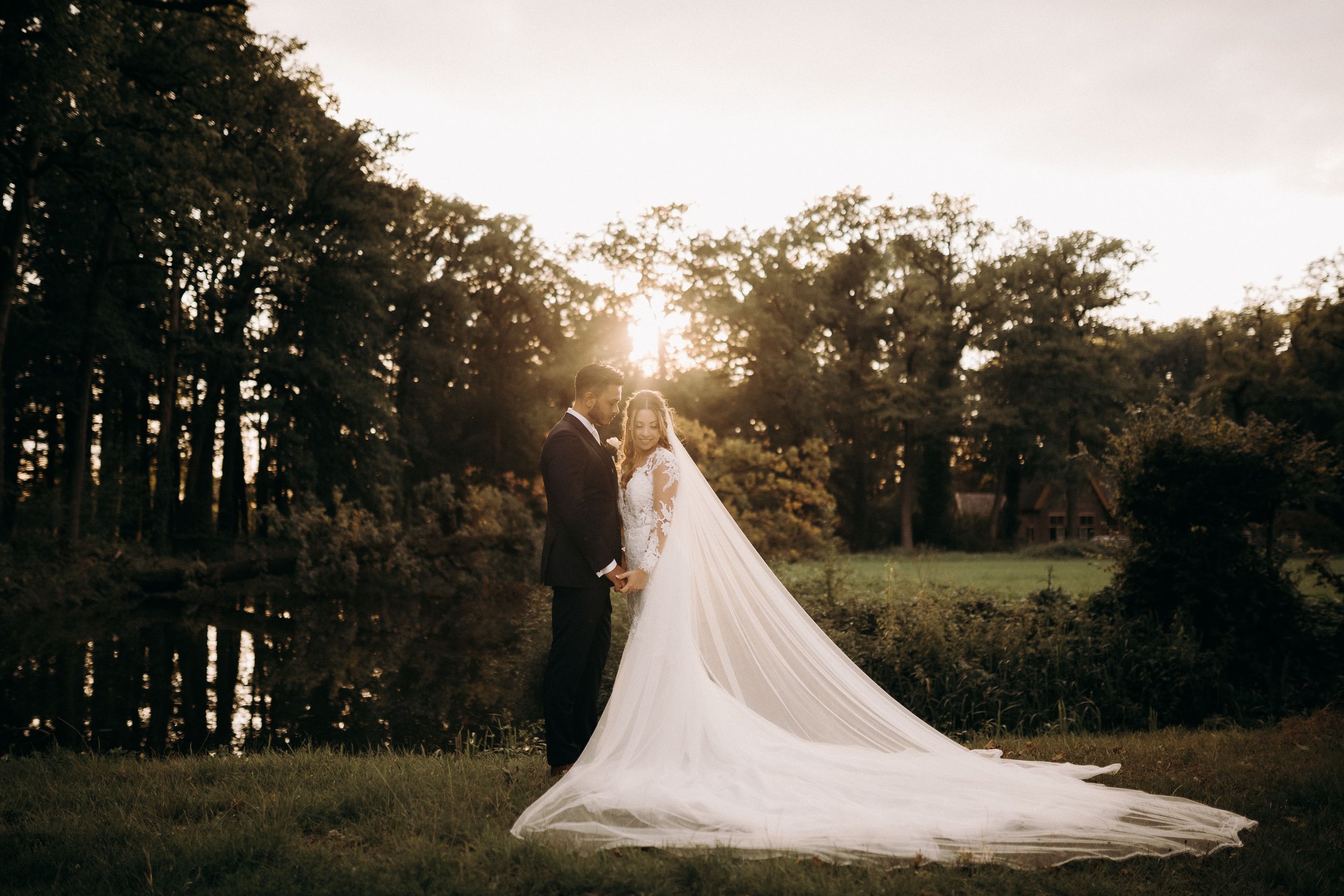 Bride and groom standing by a lakeside at sunset with the bride’s veil flowing behind her at Kasteel Heeswijk.