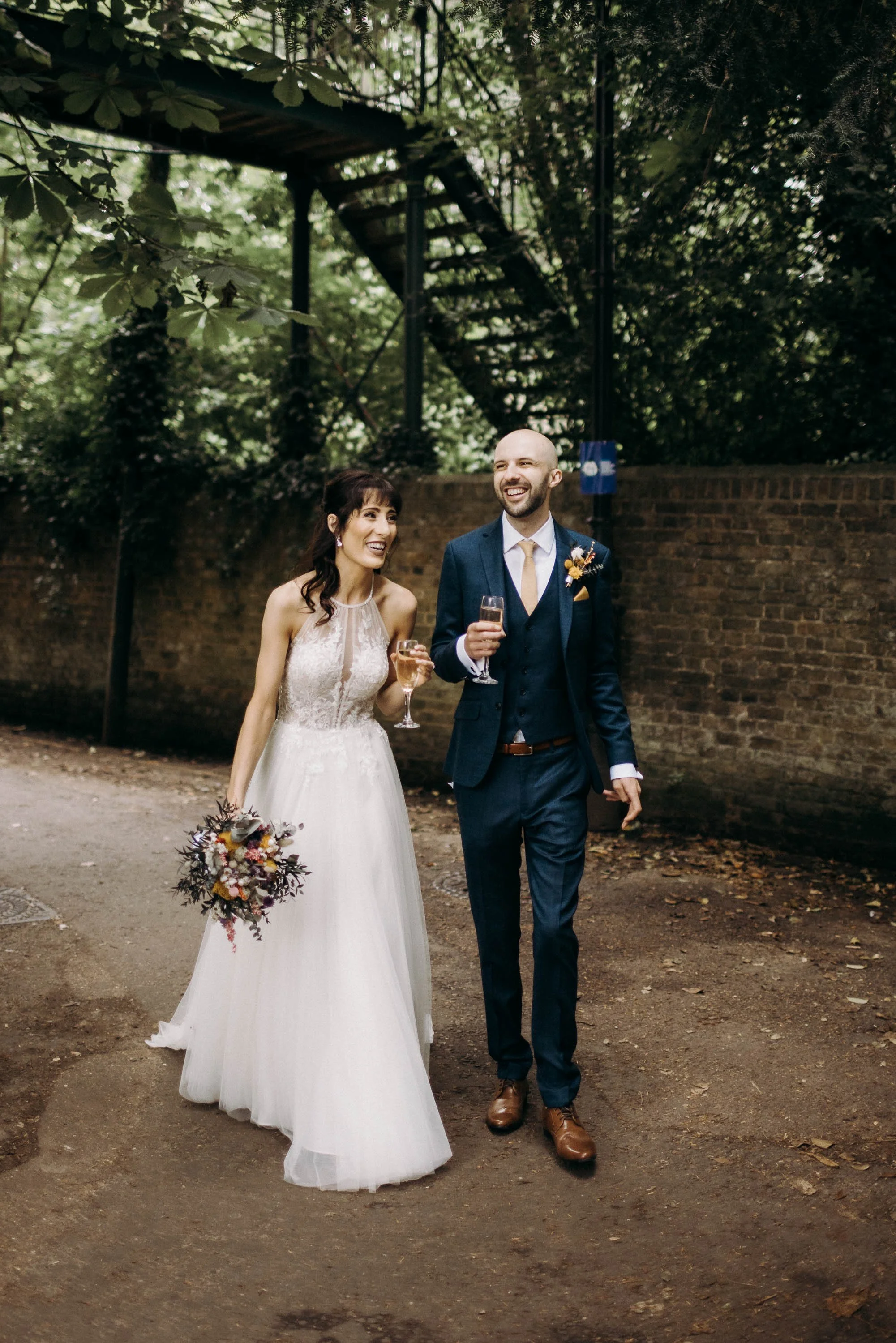 Bride and groom walking along a path beneath trees holding glasses of champagne after their ceremony at Orleans House Gallery.