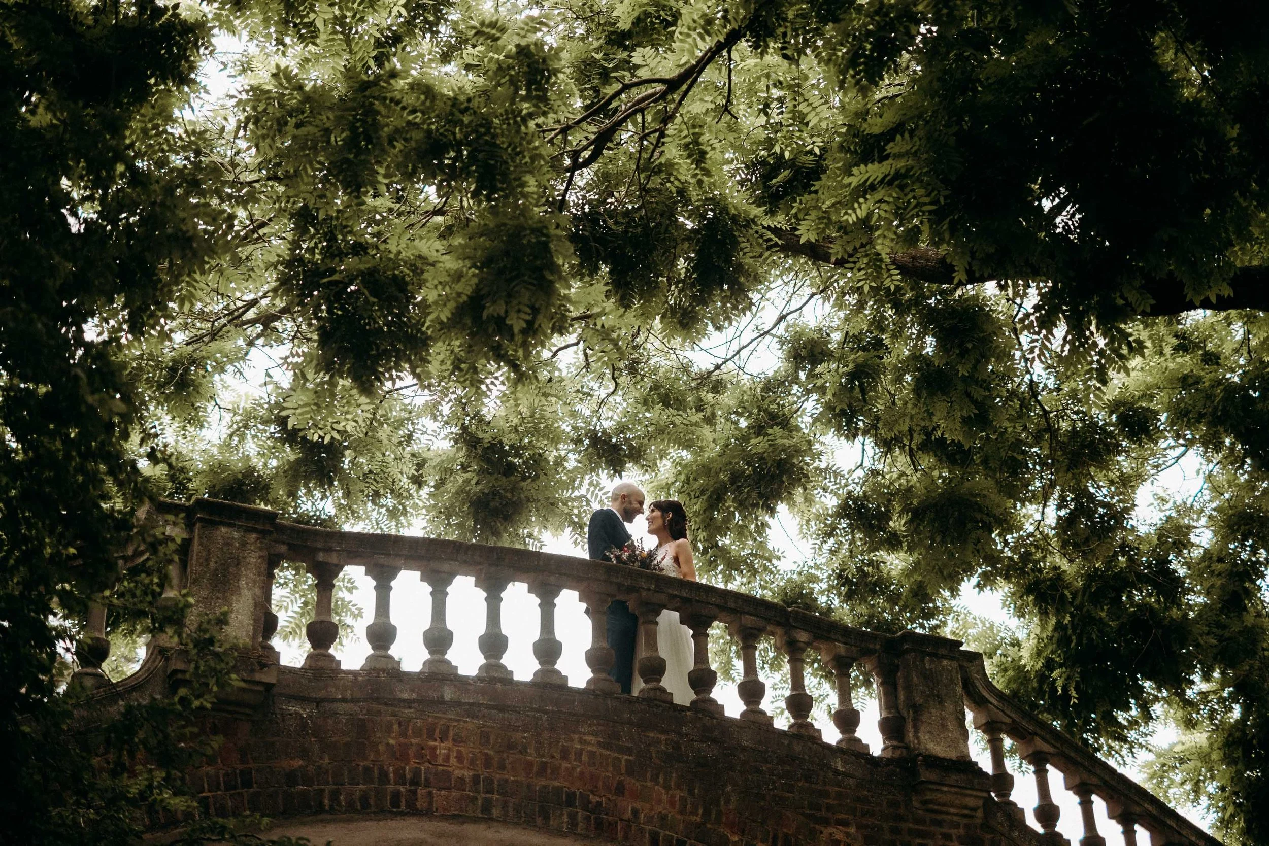 Bride and groom sharing an intimate moment on a stone bridge beneath the trees at Orleans House Gallery.
