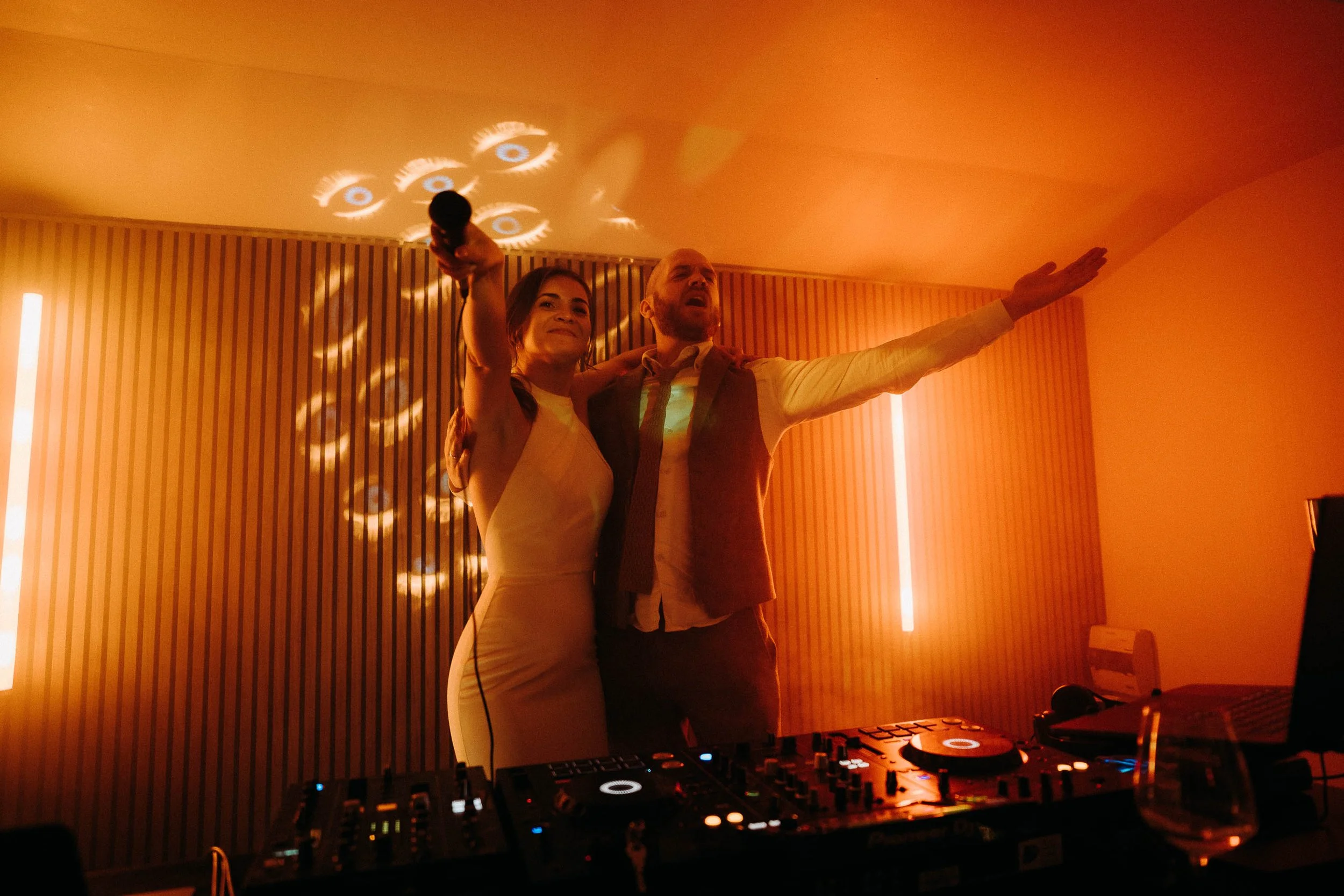 Bride and groom singing together behind the DJ booth under warm orange lighting during their wedding party.