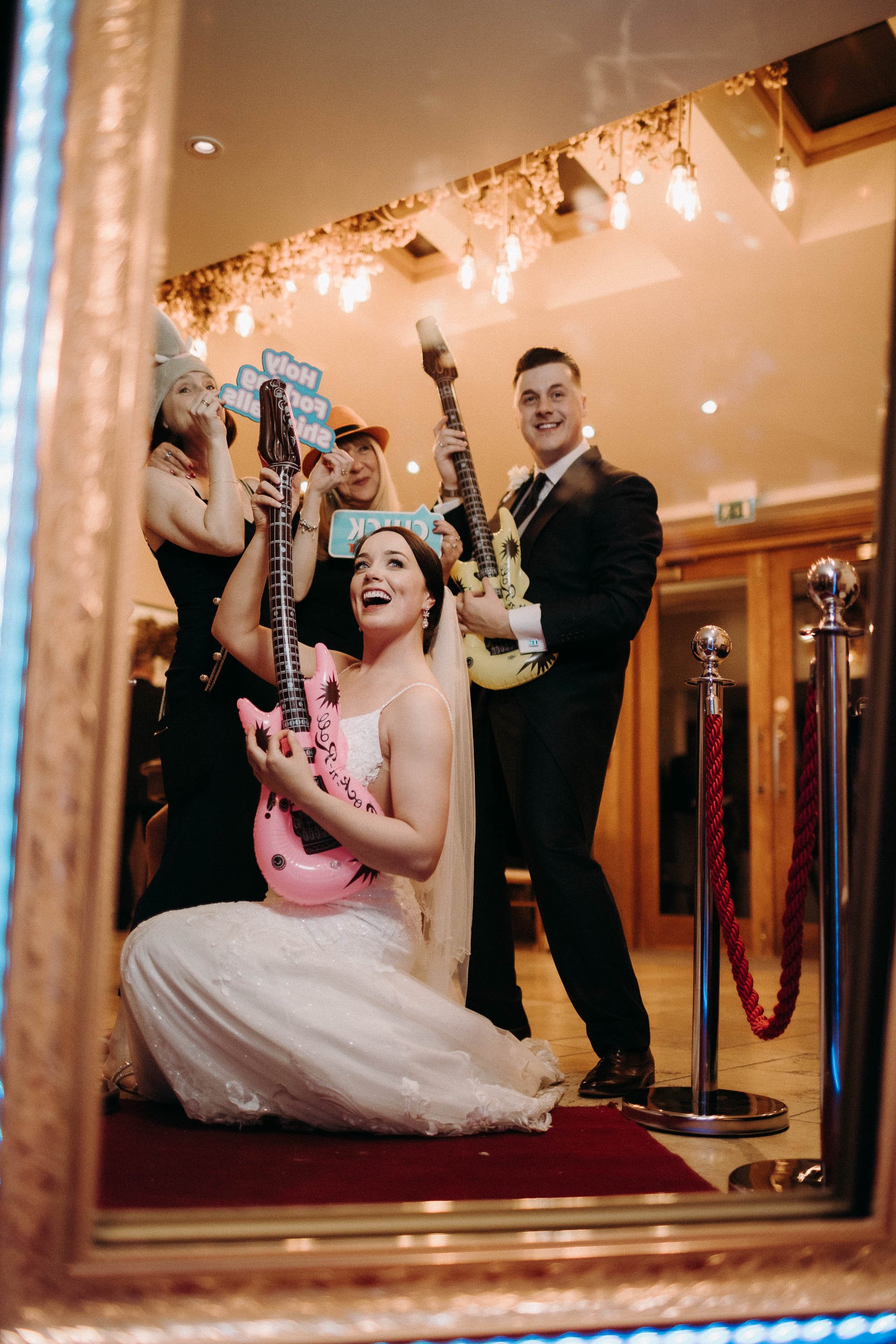 Bride and guests posing with inflatable guitars and fun props in the wedding photo booth area.