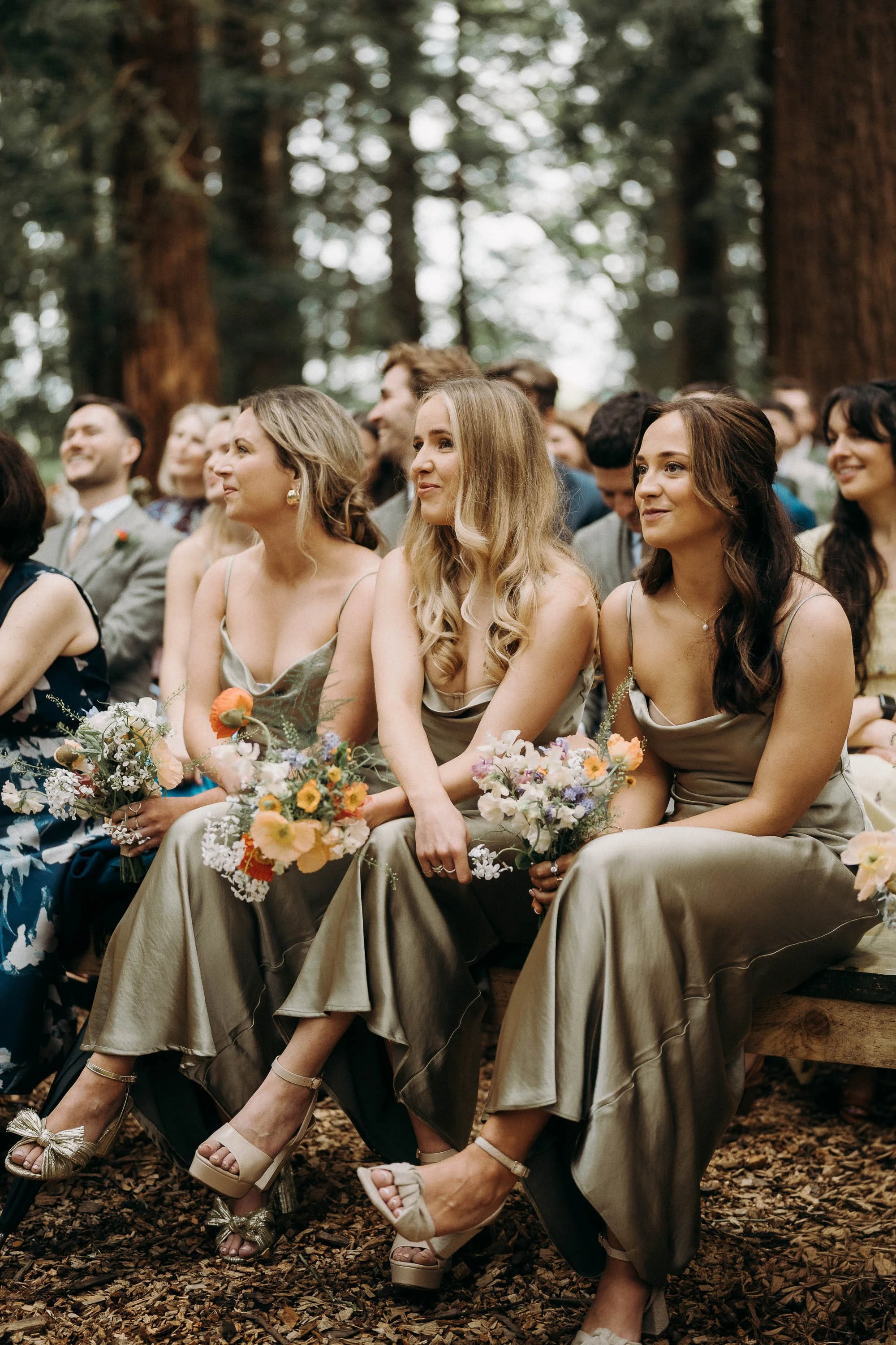Three bridesmaids in sage green dresses sit in the front row during an outdoor woodland ceremony at Two Woods Estate, holding pastel floral bouquets with tall redwood trees in the background