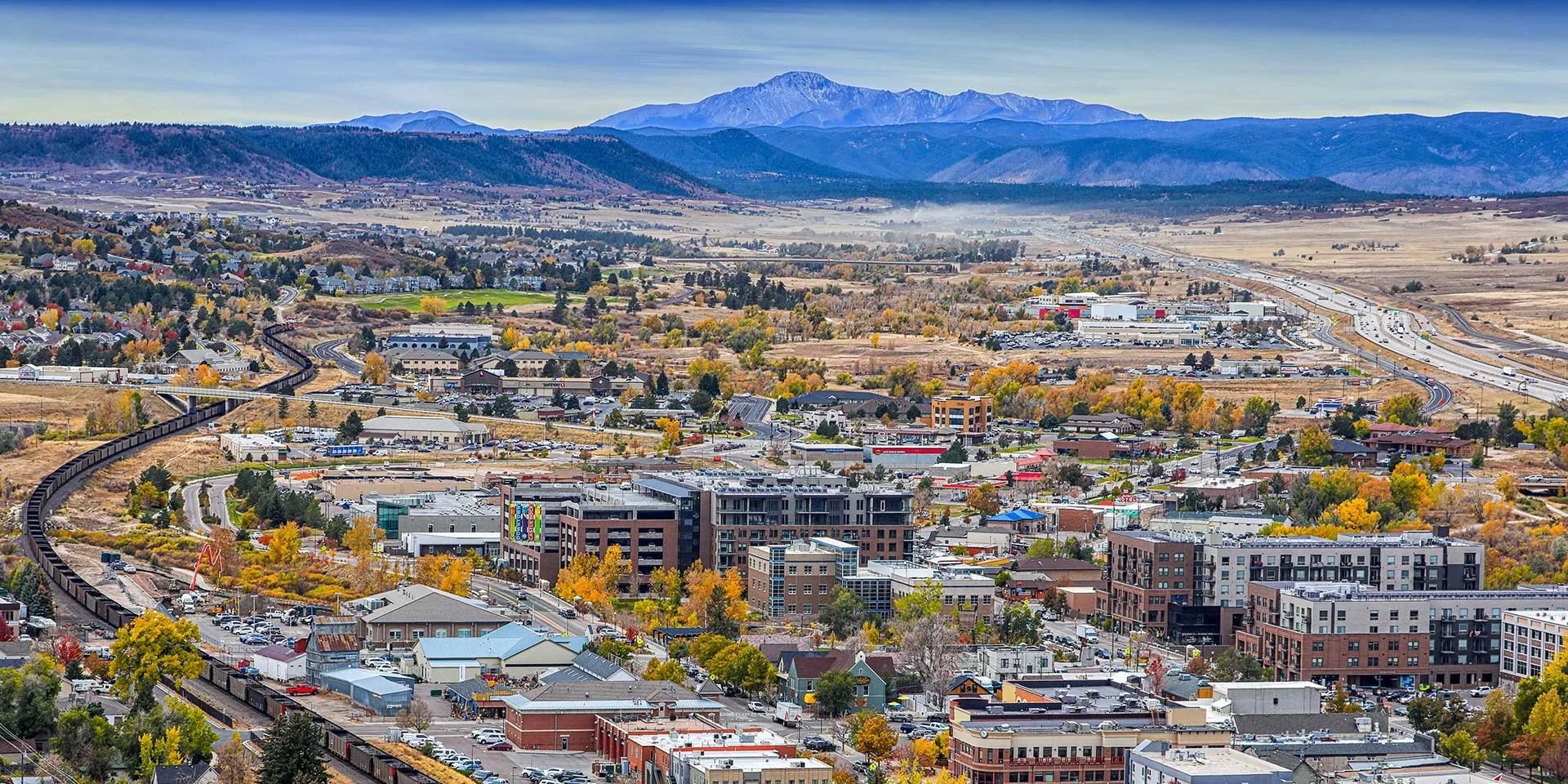 A cityscape with residential and commercial buildings, trees with autumn foliage, a mountain range in the background, and a winding train track.