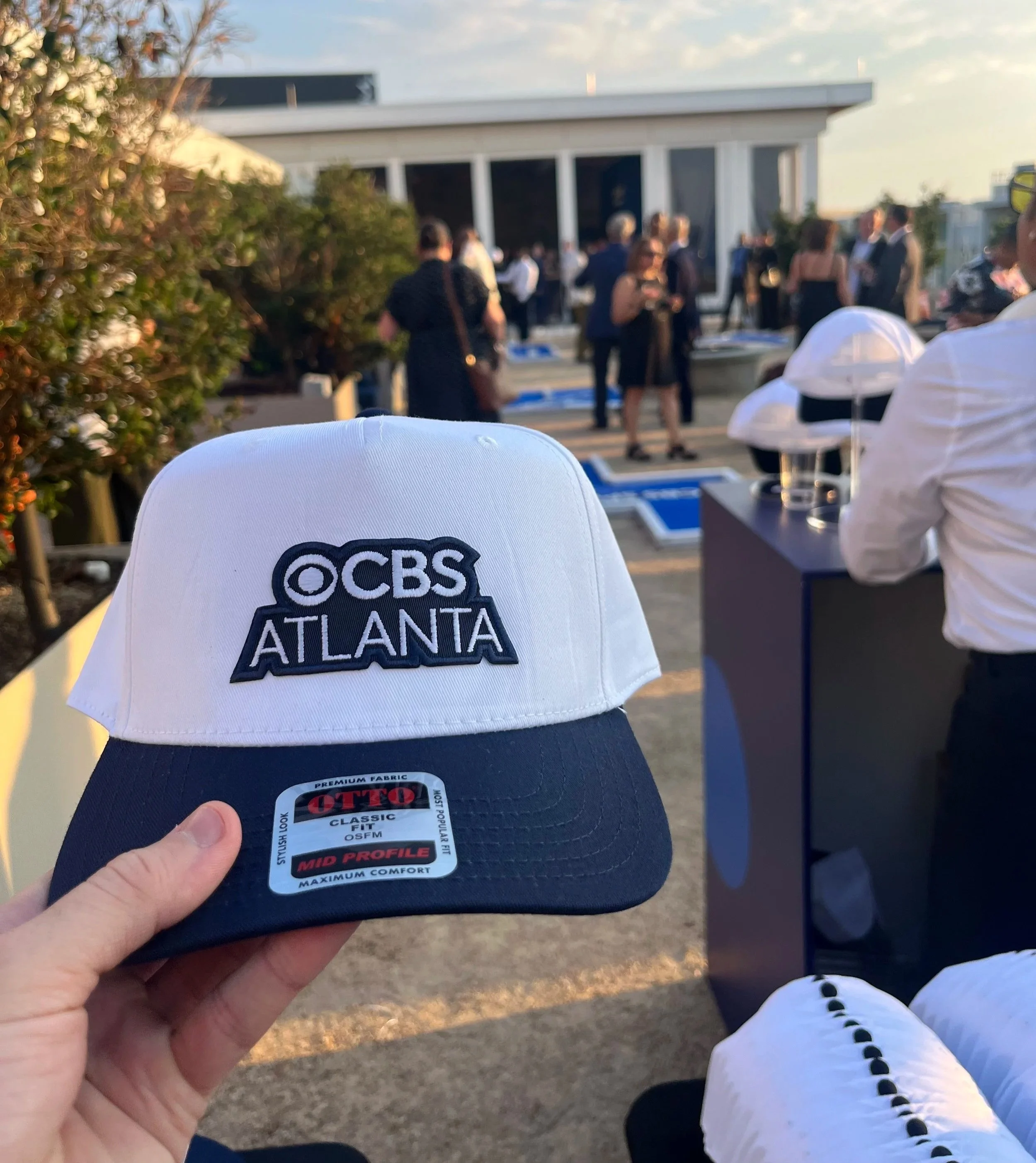 Close-up of a custom trucker hat featuring an embroidered CBS Atlanta logo patch at a live event gathering.