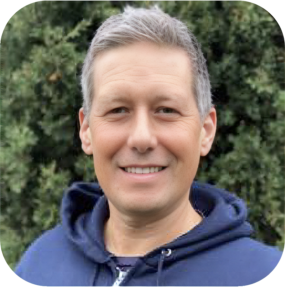 A middle-aged man with gray hair smiling outdoors with green foliage in the background.