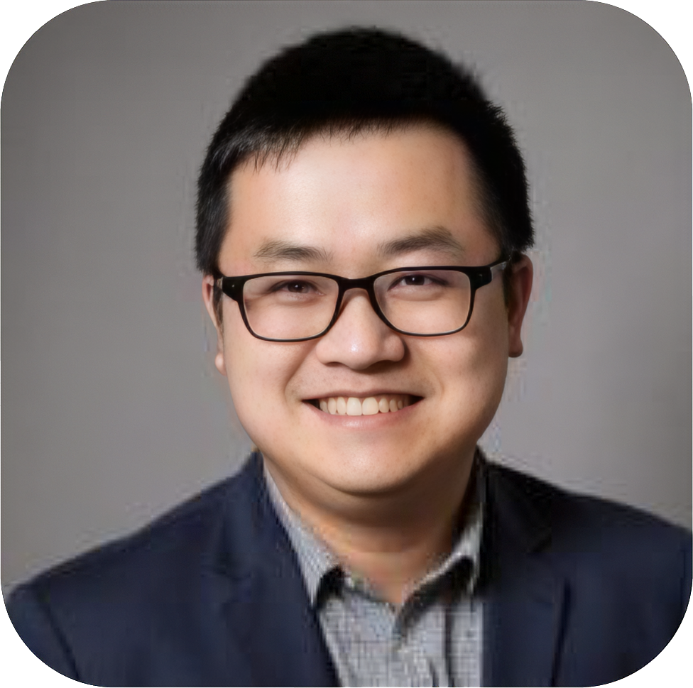 A young man with short black hair, glasses, and a broad smile, wearing a dark blazer over a collared shirt, posed against a plain gray background.