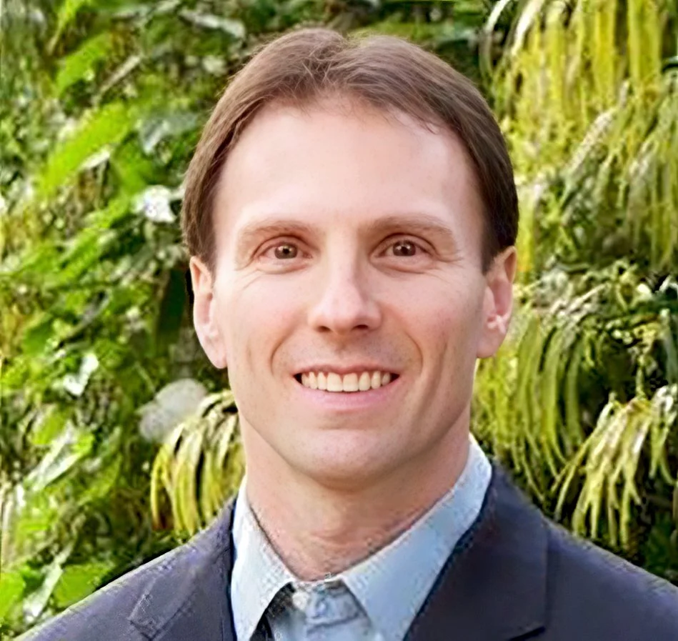 A man smiling outdoors with lush green foliage in the background.