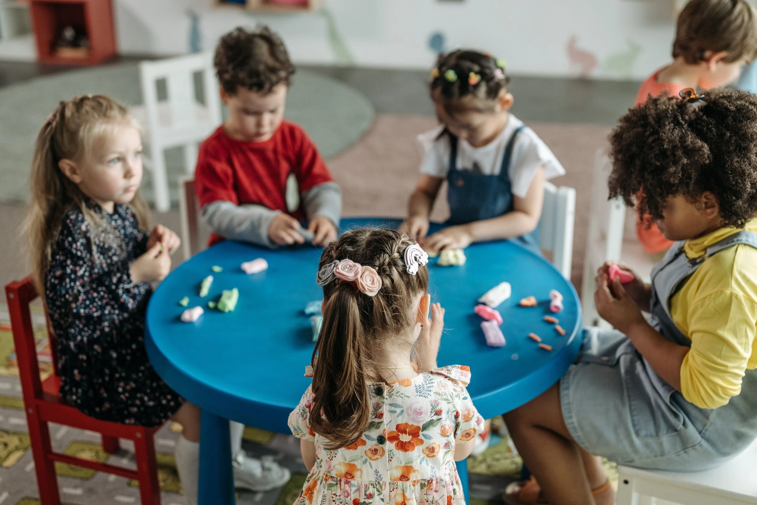 Children gathered around a blue table with colorful modeling clay, some children are sitting and some are standing, engaged in a creative activity.