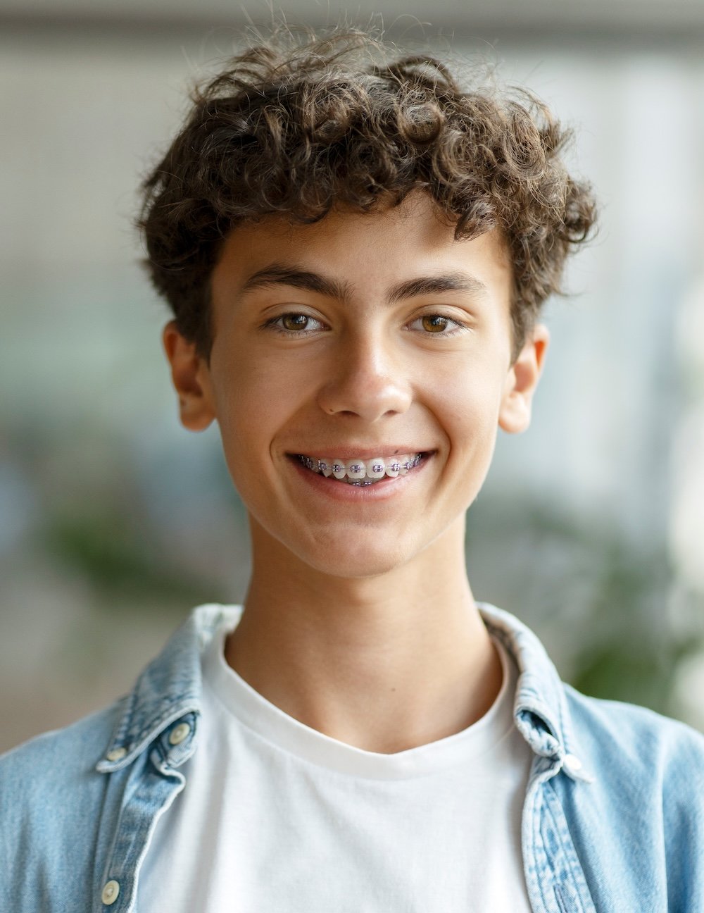 A young boy with curly brown hair and braces smiling, wearing a white t-shirt and light denim jacket, standing indoors.