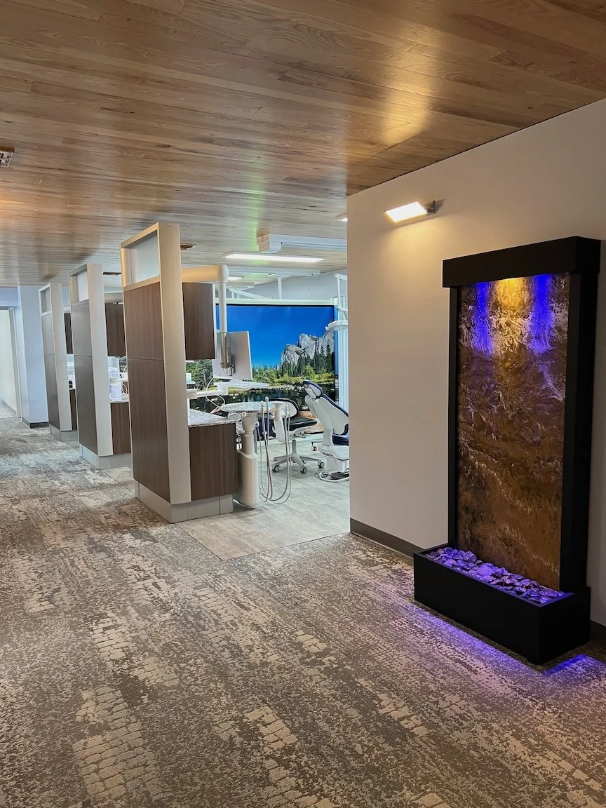 Interior view of a modern dental office with a textured water feature on the wall, blue lighting, and dental chairs in the background.