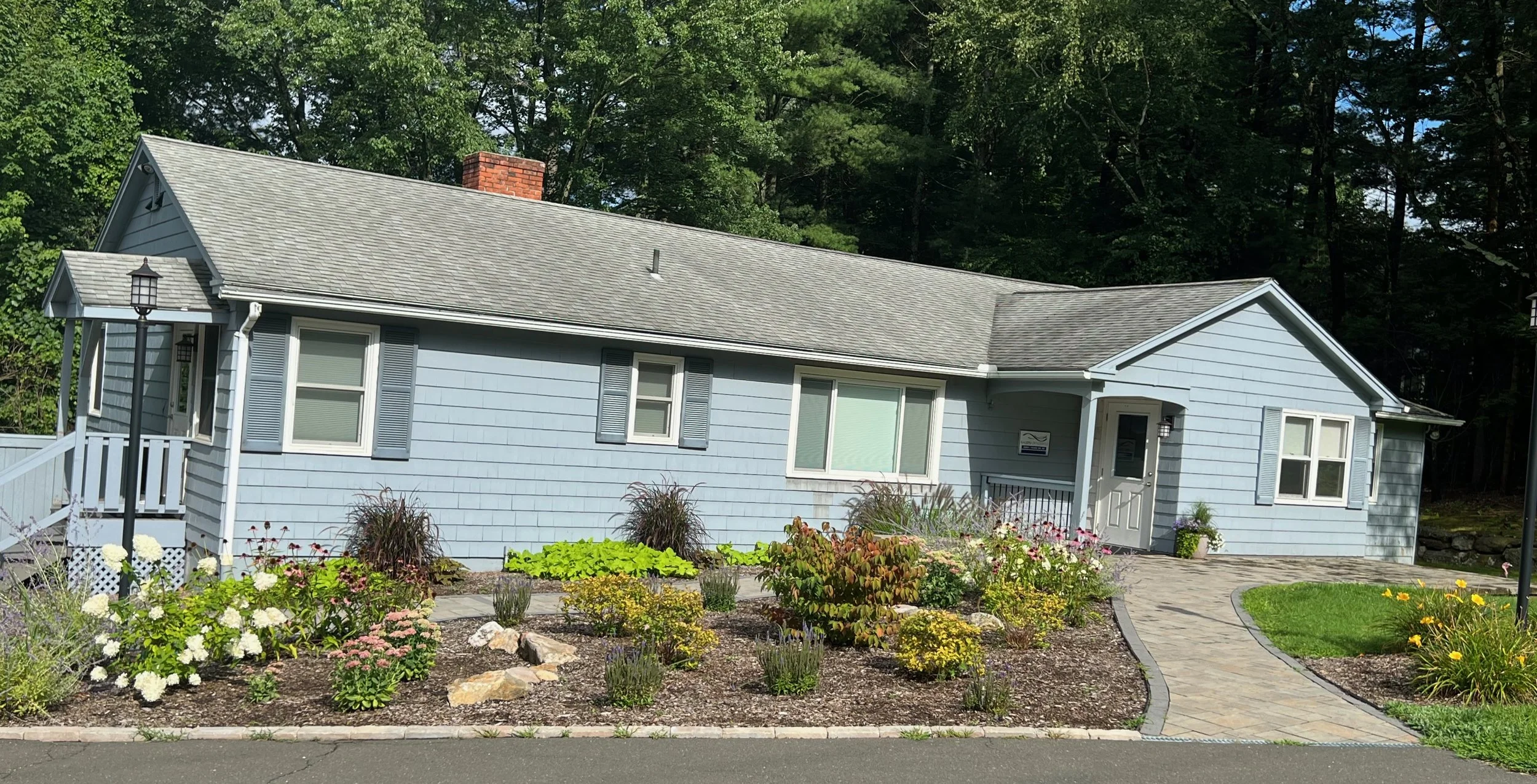 A light blue single-story house with a gray shingle roof, white door, and several windows, surrounded by a colorful flower garden and a paved walkway leading to the front entrance, with trees in the background.