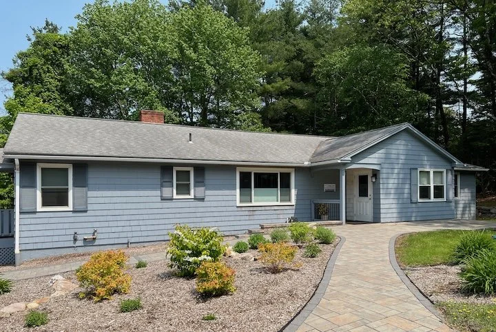 A light blue, single-story house with white trim and a gray shingled roof, surrounded by a landscaped yard with small shrubs and a brick walkway leading to the front door, set against a background of tall trees.