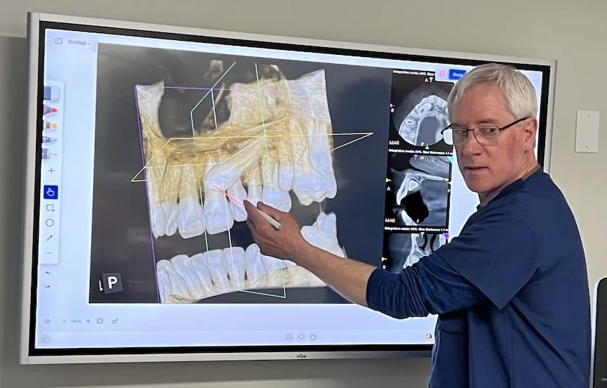 A dentist or medical professional pointing at a digital 3D model of teeth on a large interactive screen, with dental scans on the side.