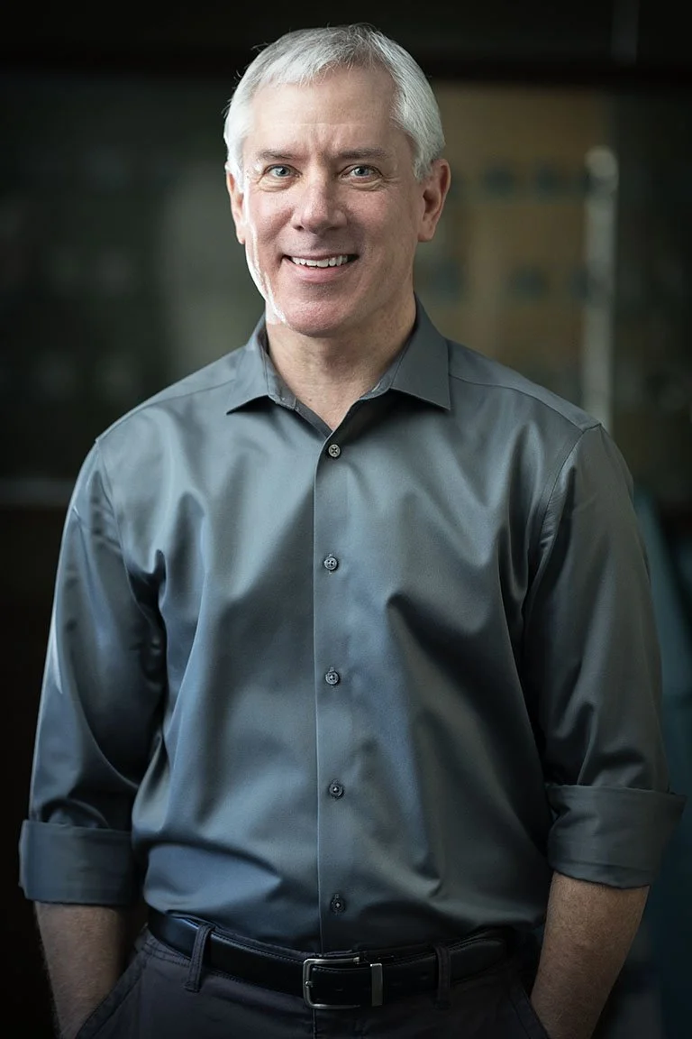 Smiling middle-aged man with gray hair in a dark gray collared shirt standing indoors.