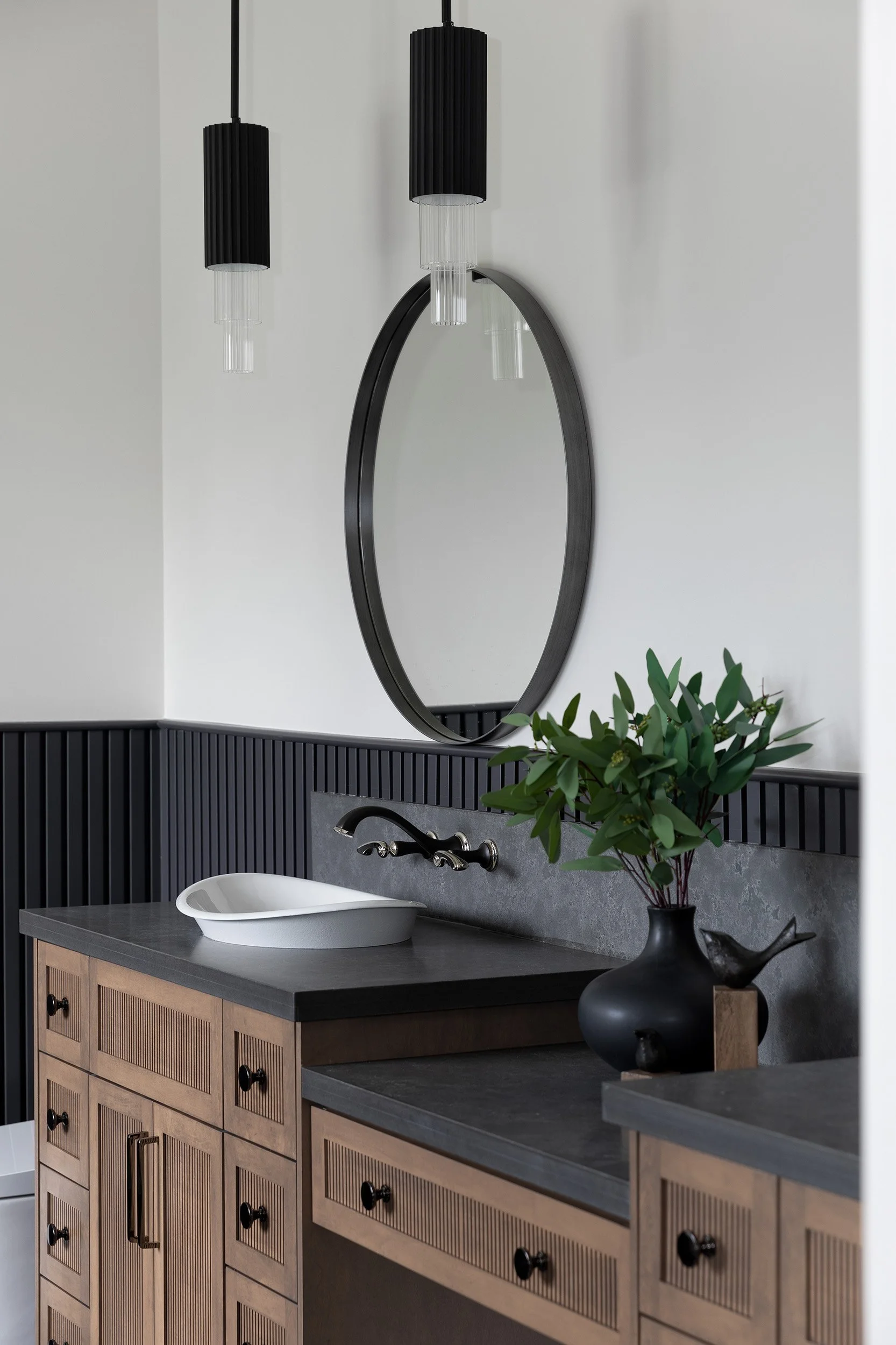 Bathroom vanity with black pendant lights, round mirror, black vase with green foliage, white sink on dark countertop, wooden cabinet.