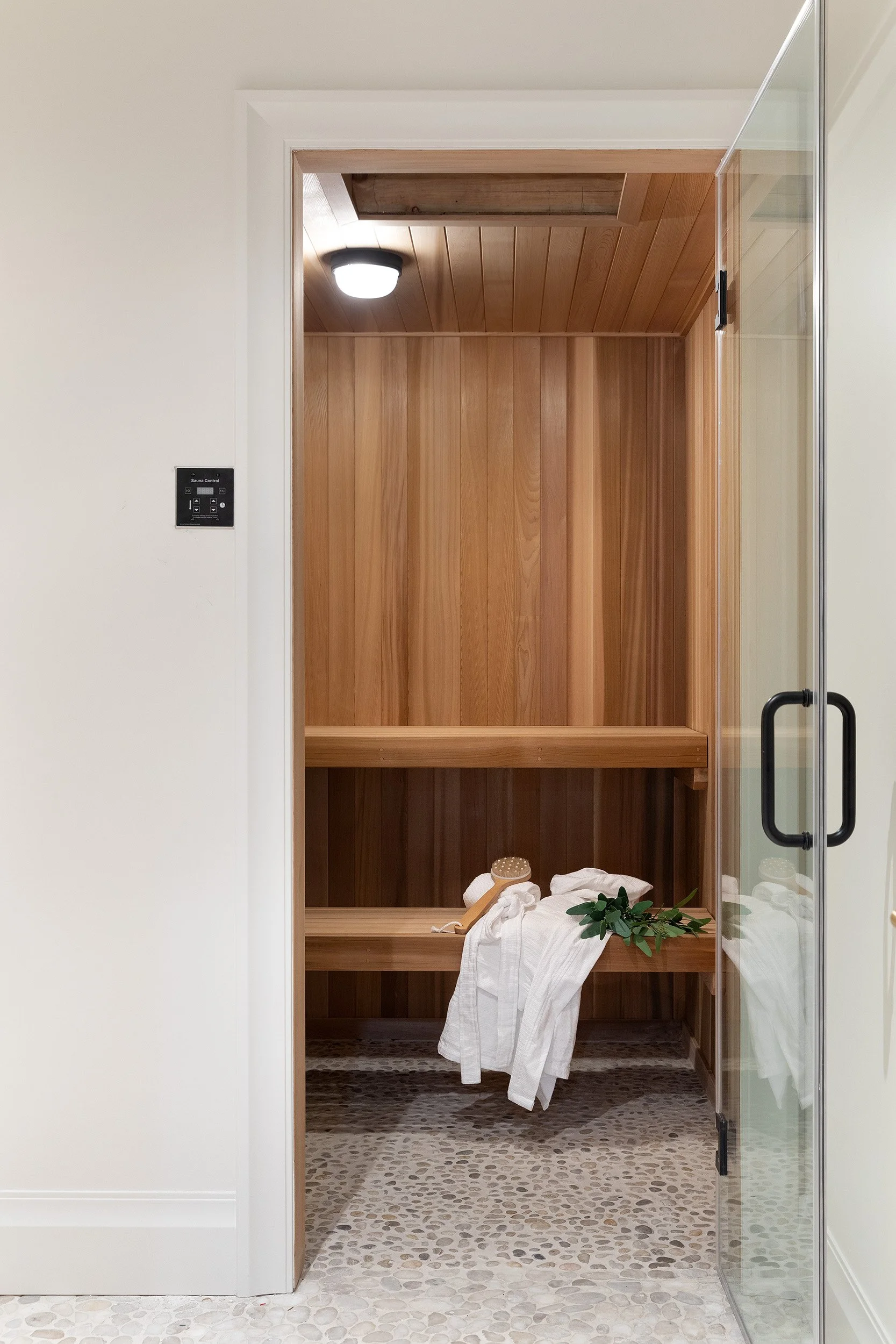 Indoor sauna room with wooden interior, glass door, white towels, a wooden sauna brush, and green leaves.