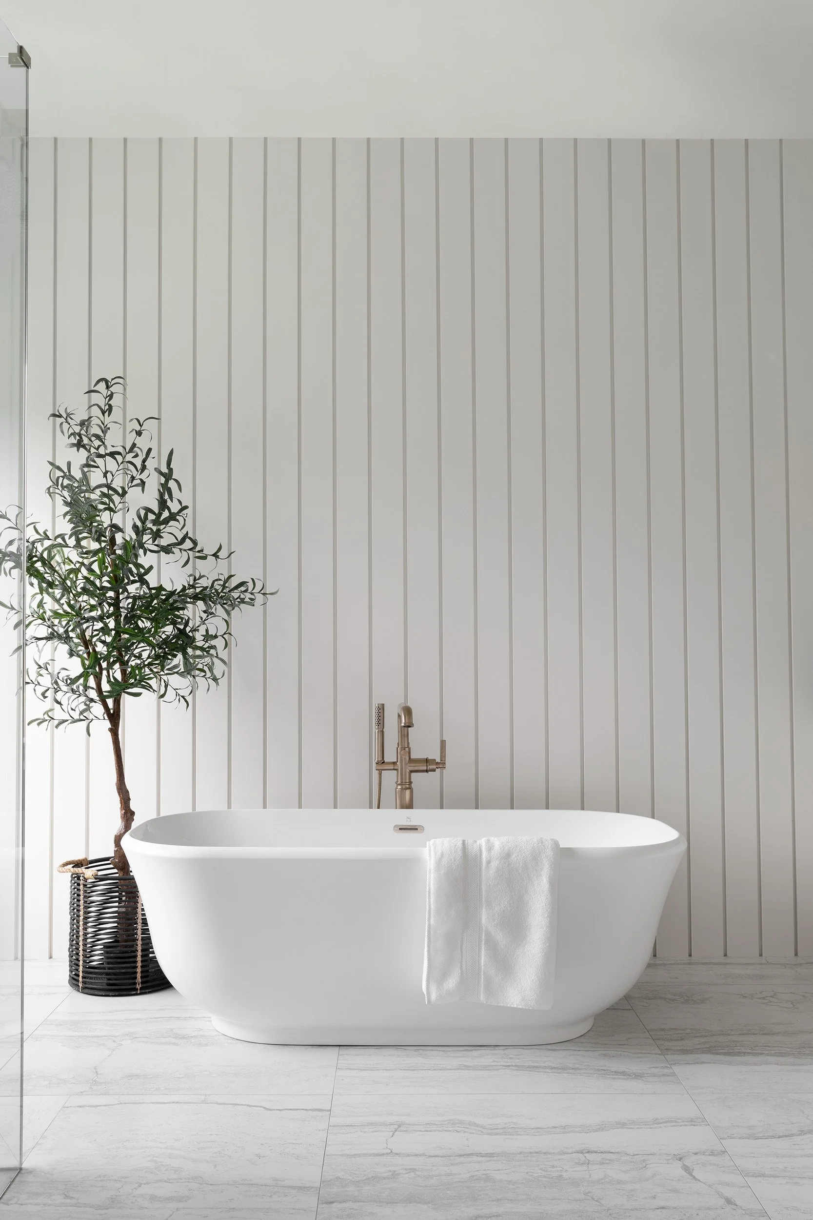 A transitional bathroom with a freestanding white bathtub, a towel draped over the side, a gold faucet, a potted plant, and a light-colored wood floor and wall paneling.