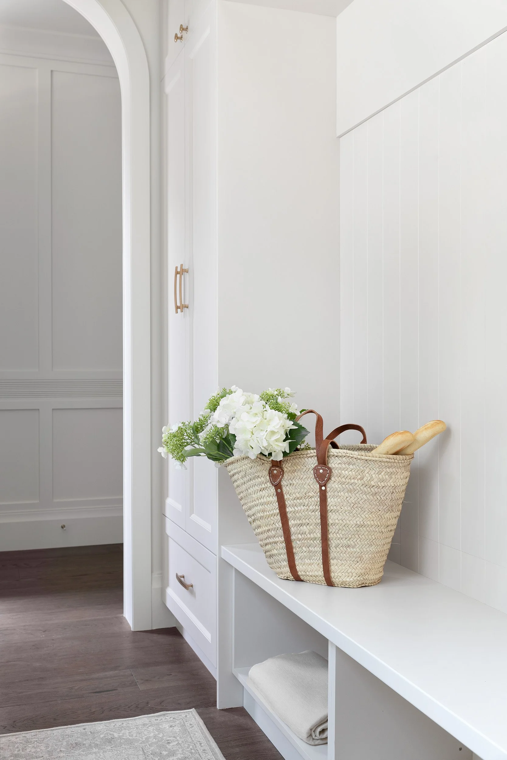 White entryway with a woven basket containing white flowers and baguettes on a built-in bench, beige rug below, wood flooring.