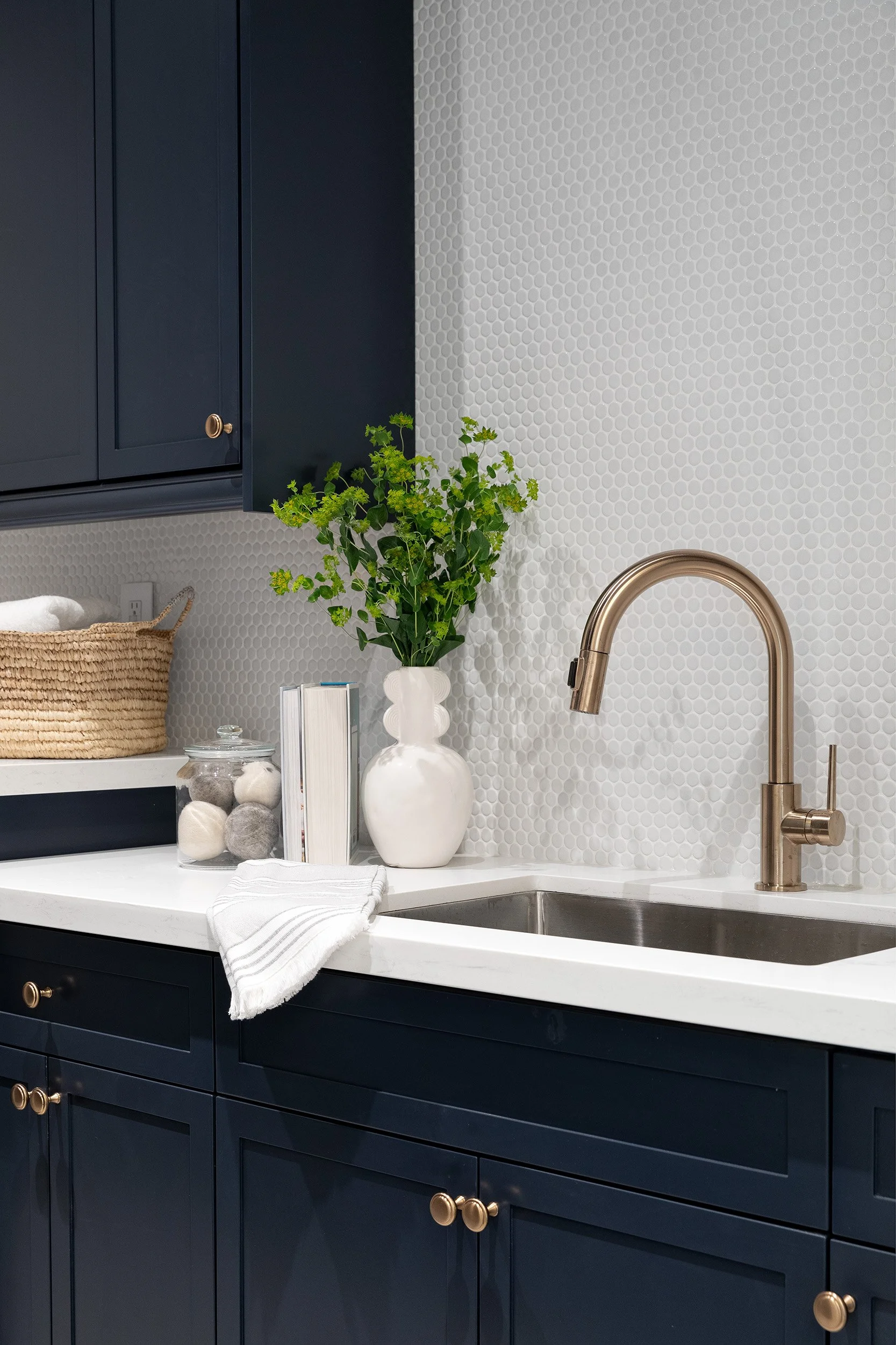 Laundry room with navy blue cabinets, a white countertop, and a brass faucet. A white vase with green foliage, a glass jar with felt balls, and a white dish towel are on the counter. The wall behind has small white penny tiles.