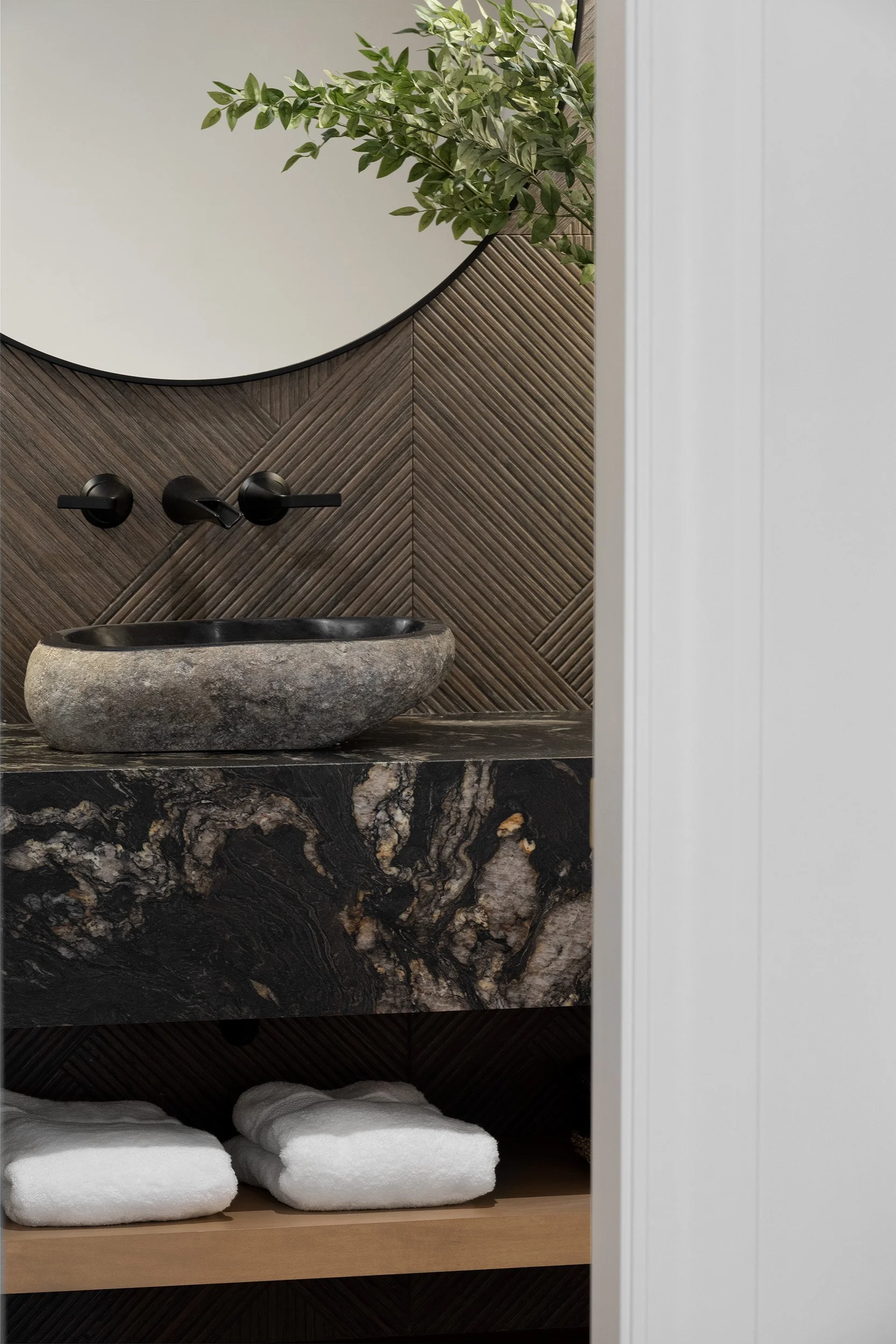 A modern bathroom vanity with a black and gray stone sink on a dark marble countertop, white towels below, a round mirror above, a wooden floating shelf with greenery and patterned  wood wall tile.