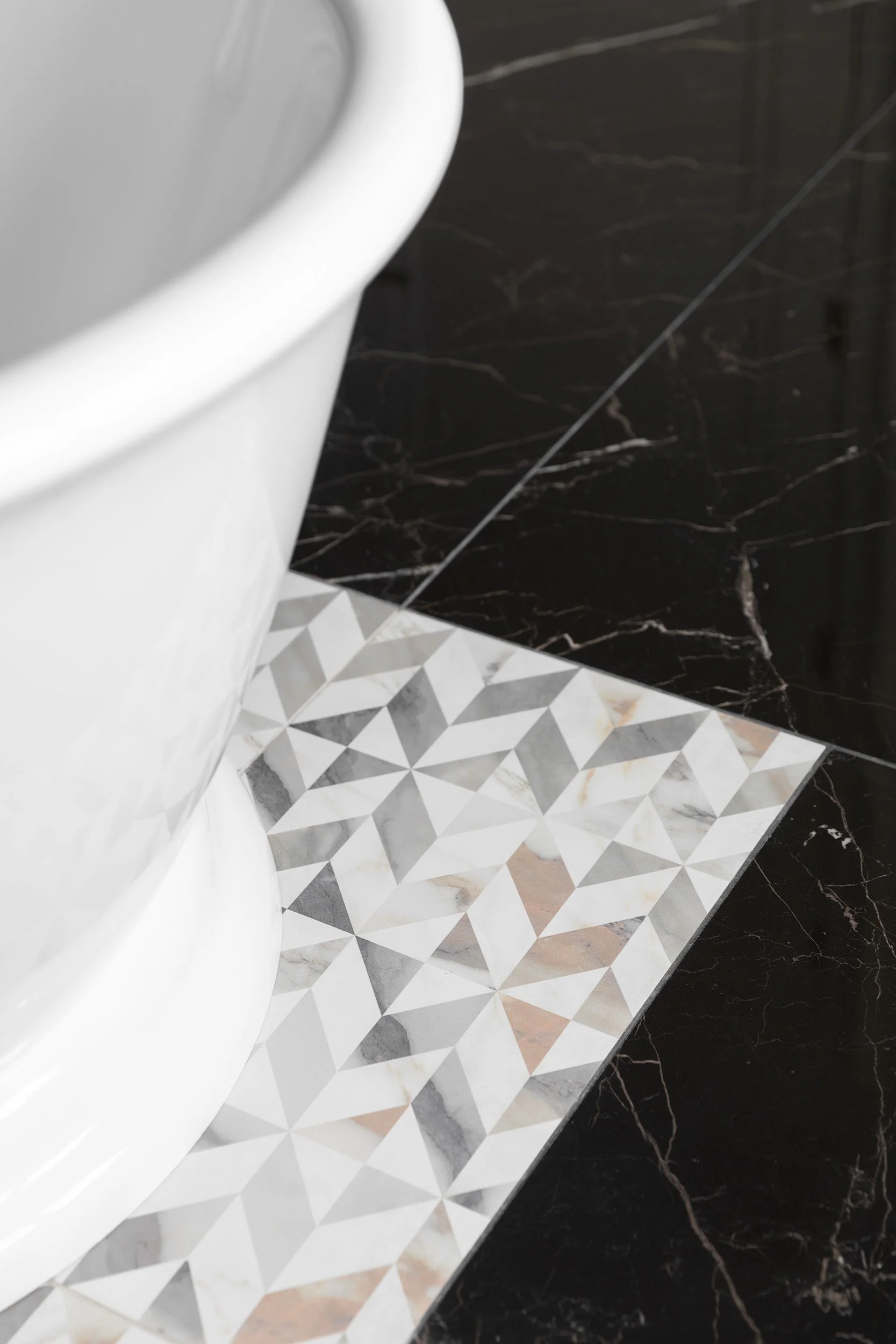 Close-up of a bathroom floor showing a section of patterned white and gray tiles adjacent to black marble tiles. Part of a freestanding tub is also visible.