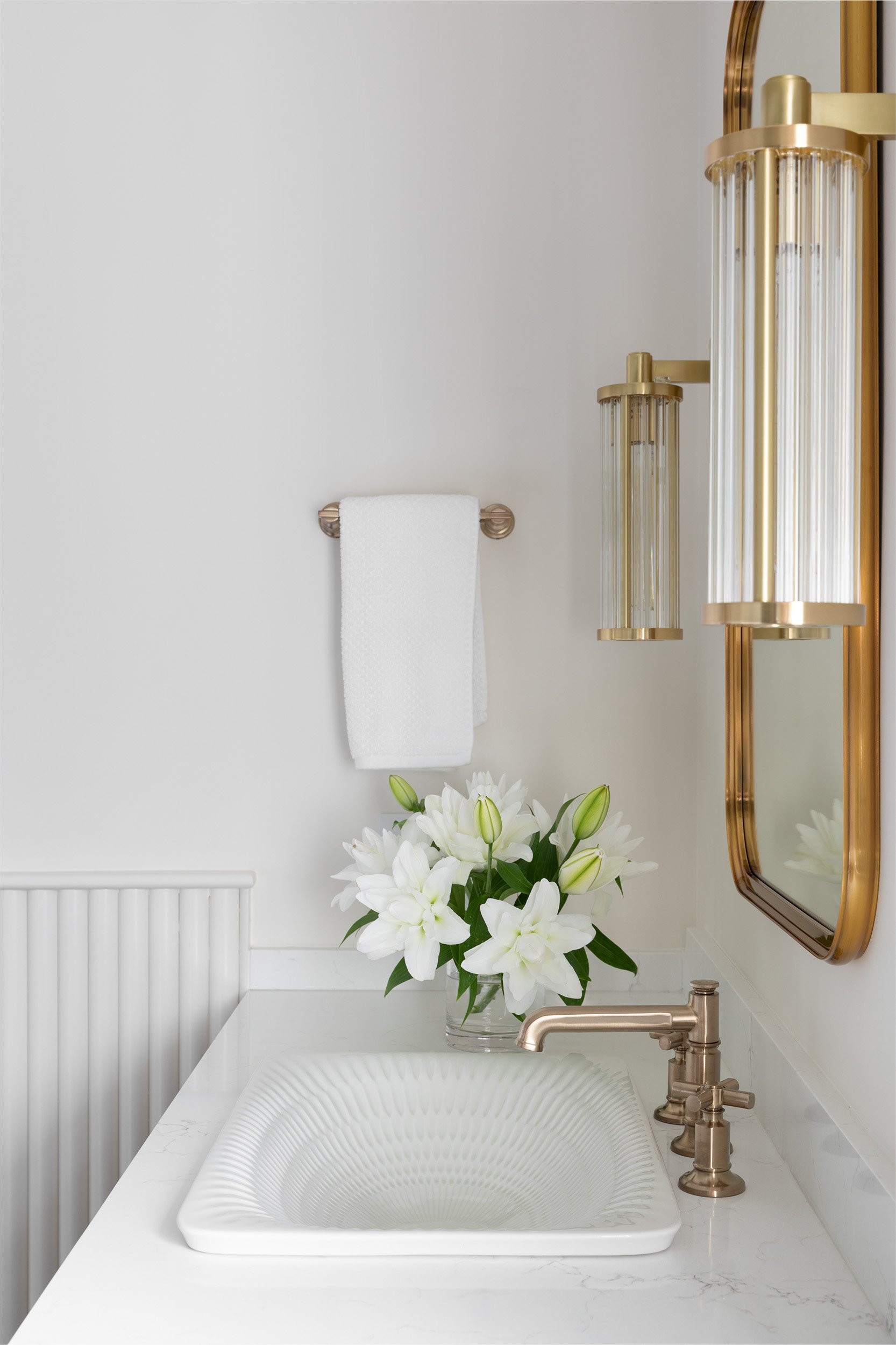 A bathroom sink with a white floral arrangement in a glass vase, a white hand towel on a wall hook, and transitional gold fixtures including a mirror, sconces, and a faucet, with a white marble countertop and paneled wall.