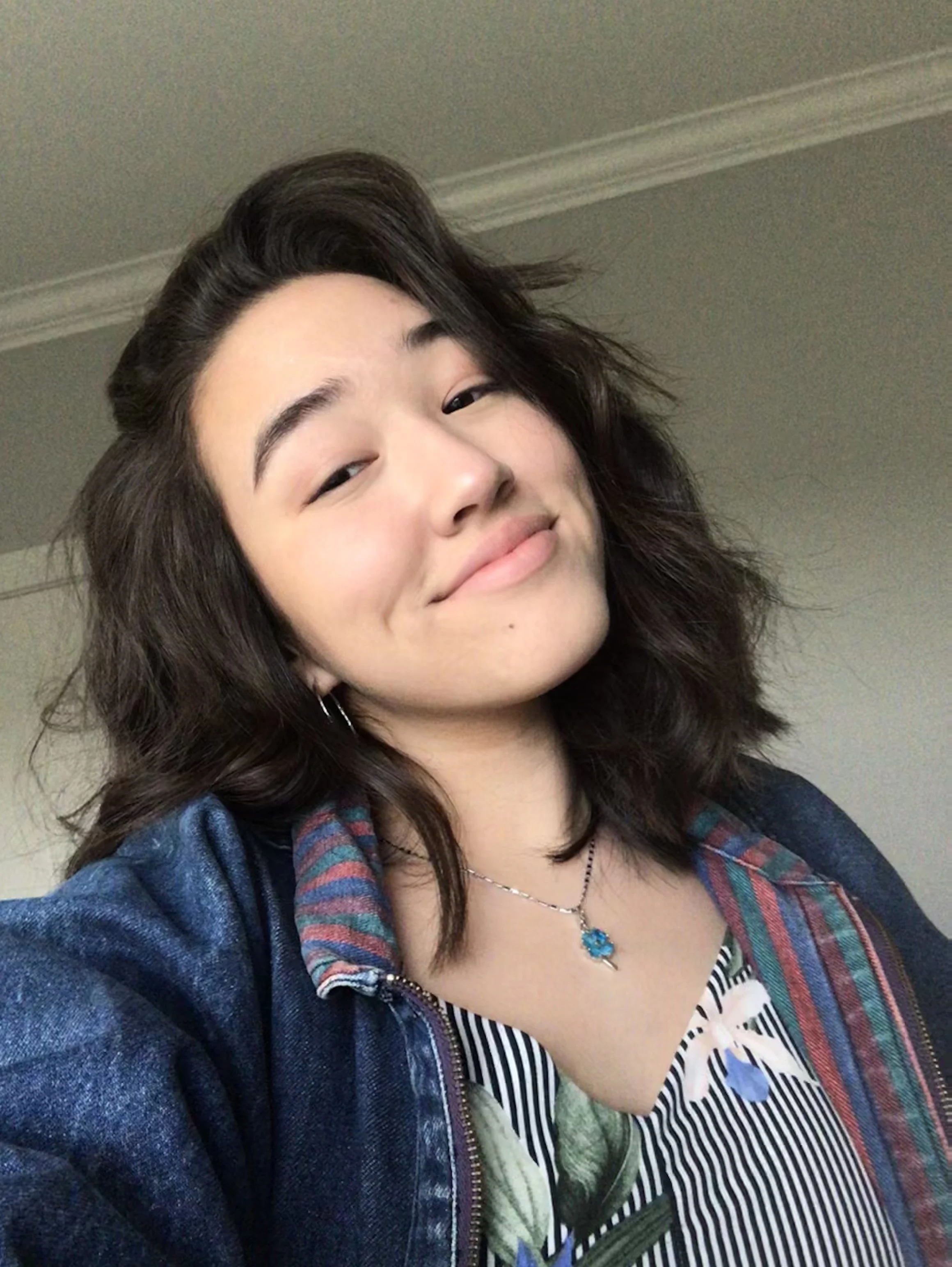 Selfie of woman in her 20's, head tilted to the left, shoulder length brown hair and a closed mouth smile. Wearing a jean jacket and collared shirt with a pendant with a green stone.