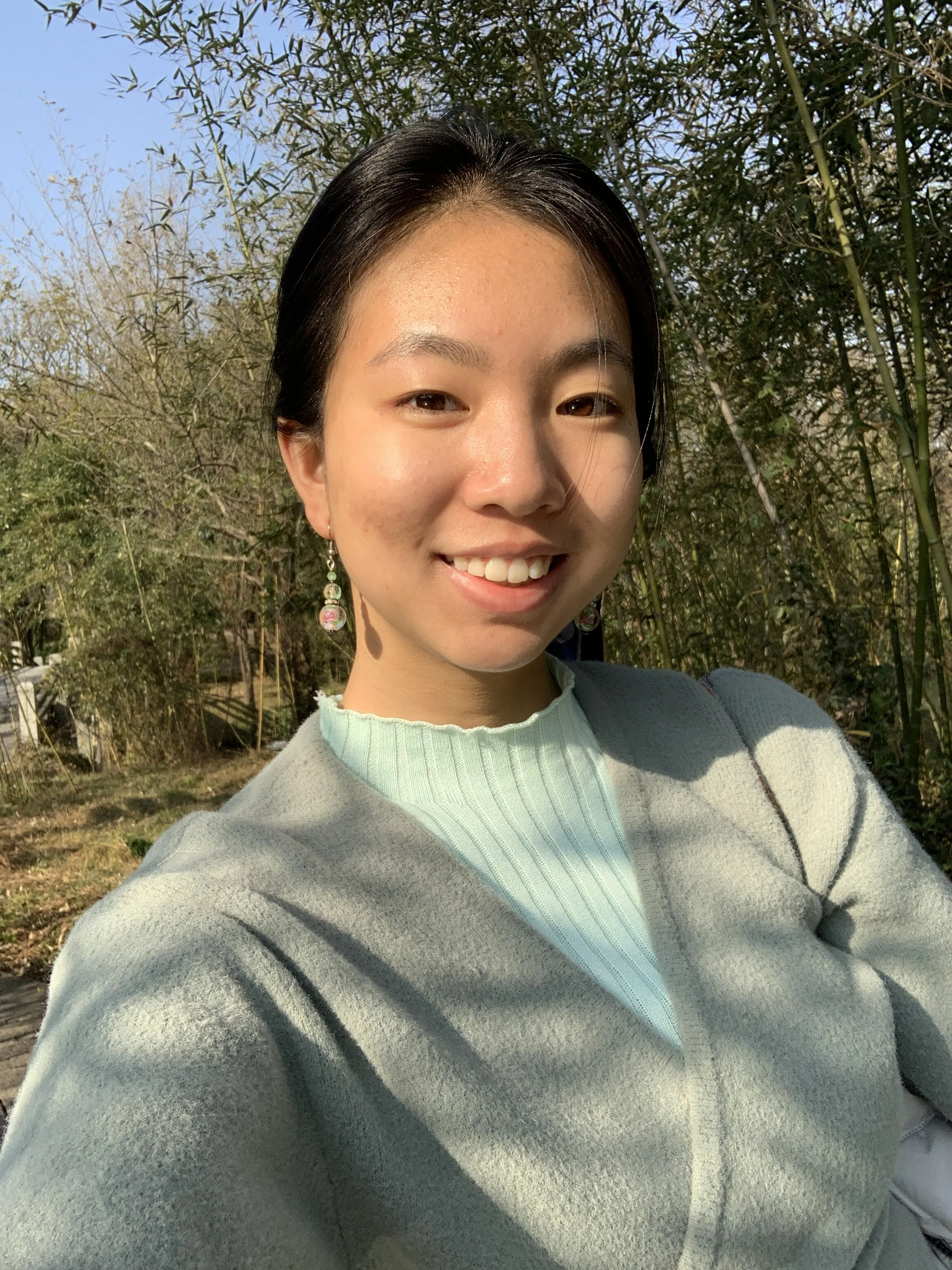 Picture of young Asian woman wearing teal sweater and gray cardigan, in front of trees, hair pulled back and smiling. The sun is shining.