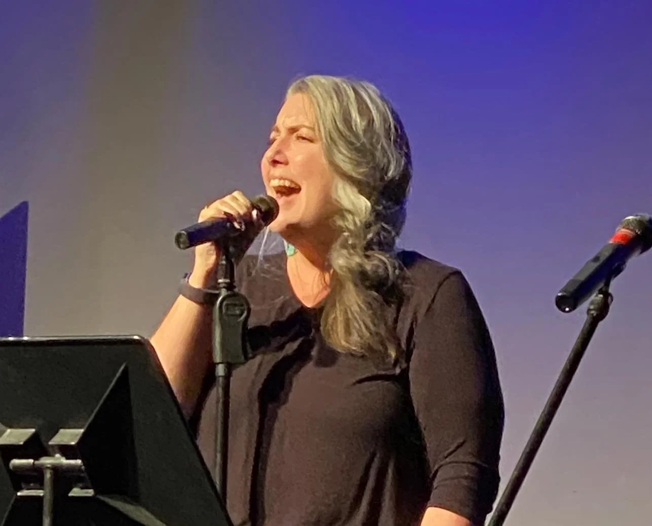 Picture of Joslin in front of a microphone belting out a high note, hair in a side braid and wearing a black shirt