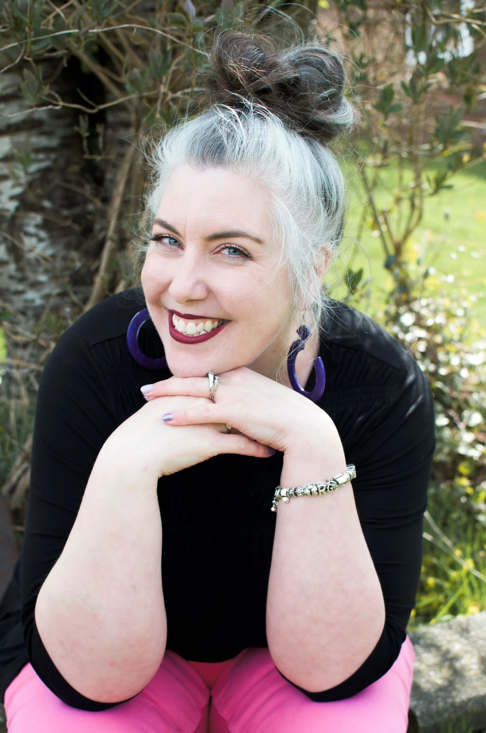 Joslin sitting in front of a tree wearing her hair in a bun, black top with pink crop pants, smiling into the camera. Charm bracelet on left wrist and purple hoop style earrings.