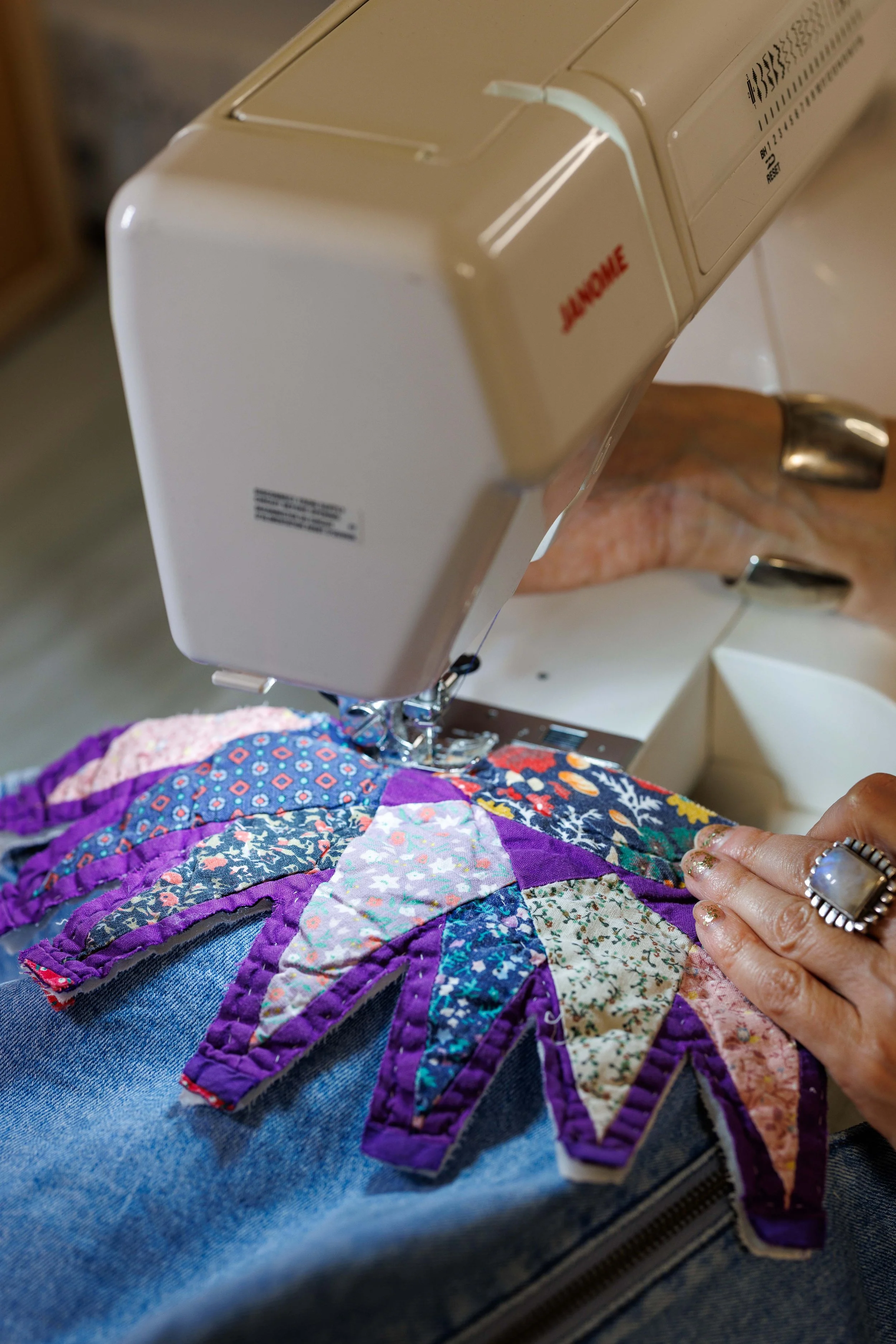 Close-up of a person sewing a colorful quilt on a sewing machine. The person's hand is guiding the fabric quilt, which has a vibrant patchwork pattern, and they are wearing a ring and nail polish.