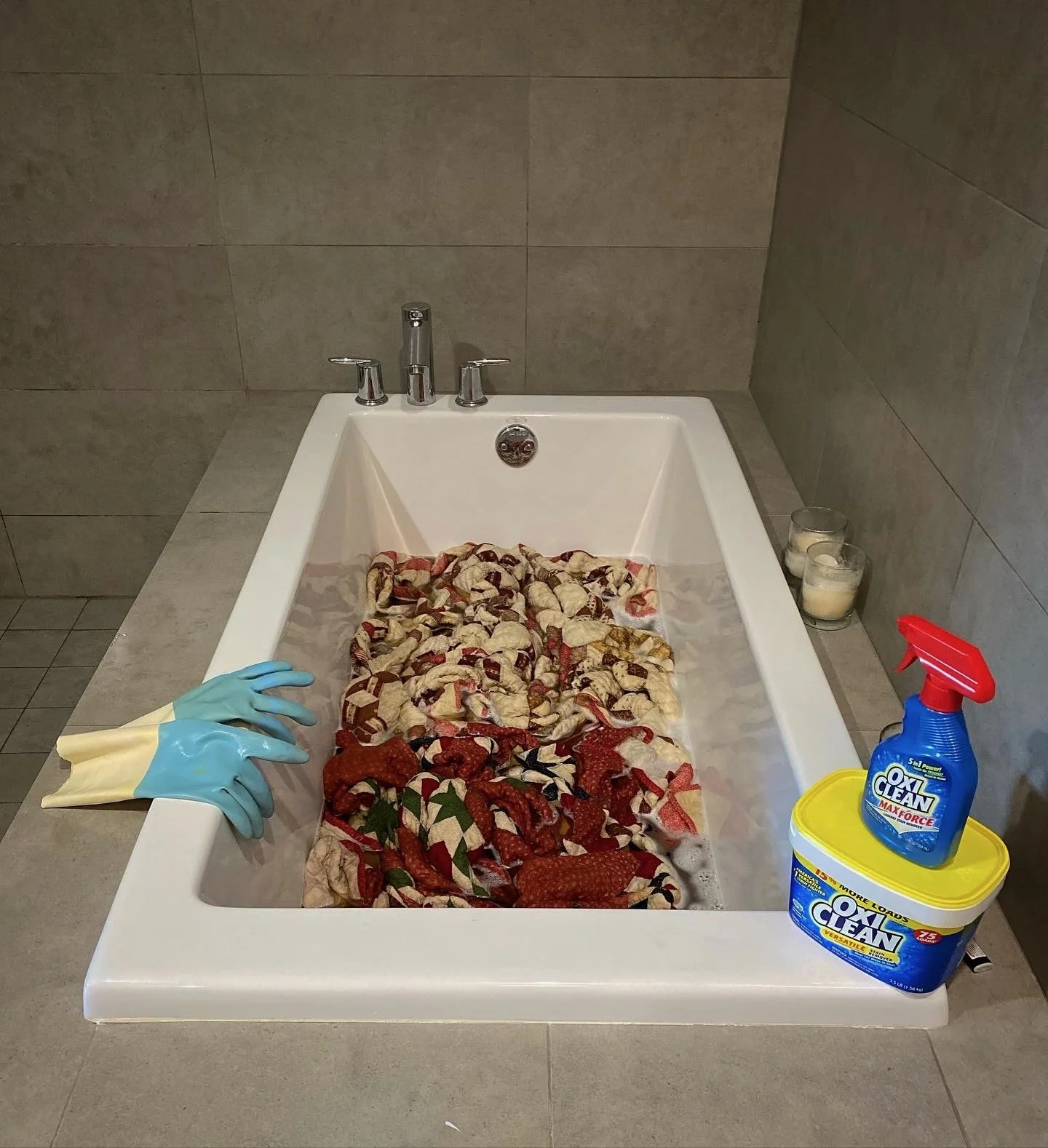 A bathroom with a bathtub filled with holiday-themed fabric or clothing, cleaning supplies including Oxiclean on a table, candles on the side, and a pair of rubber gloves resting on the edge of the bathtub.