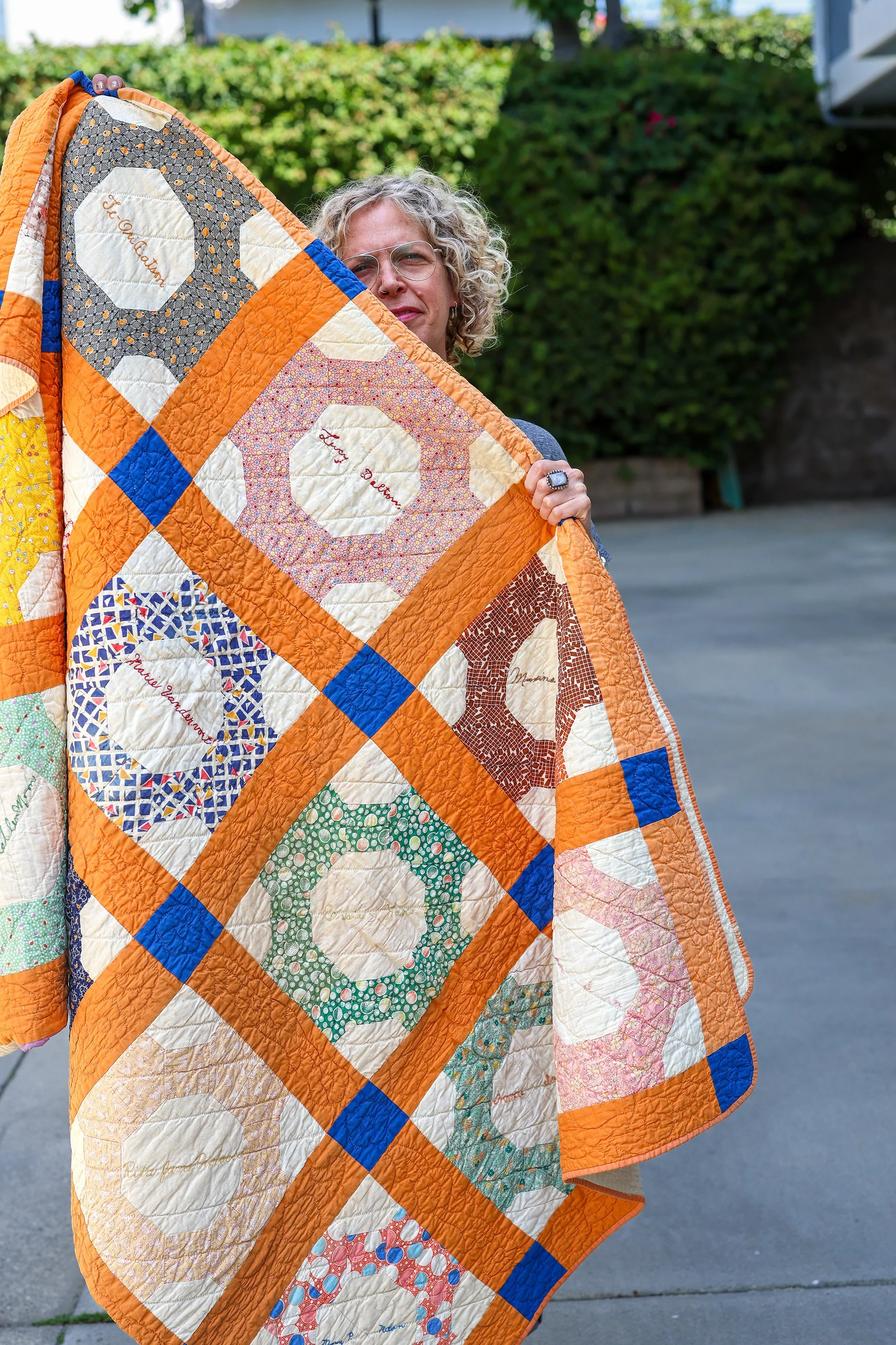 Person holding a colorful quilt with various patterns and words embroidered on some patches, standing outdoors with greenery and a paved surface in the background.