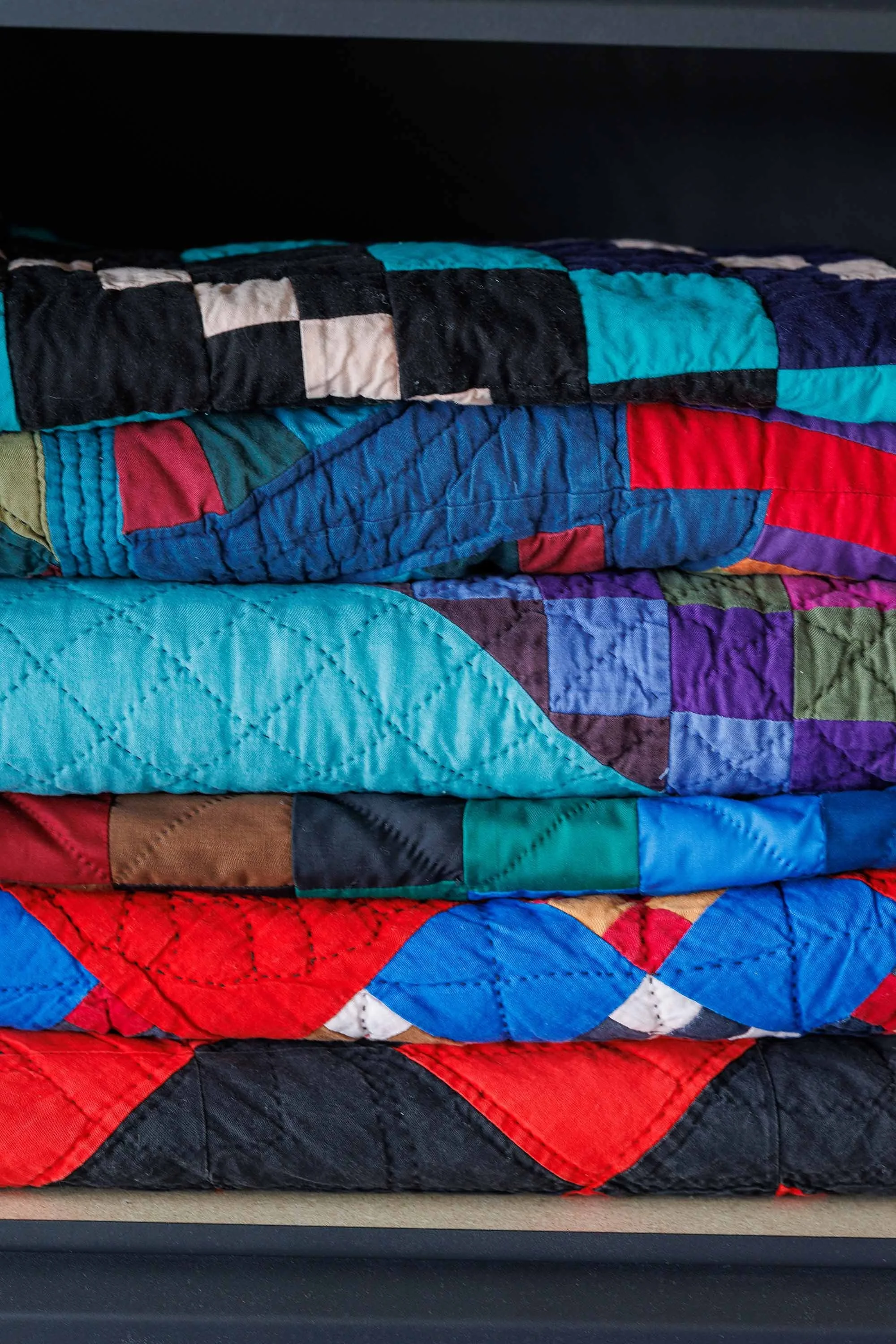 Stacked colorful patchwork quilts featuring various geometric patterns and bright colors such as red, blue, purple, and black, resting on a surface.
