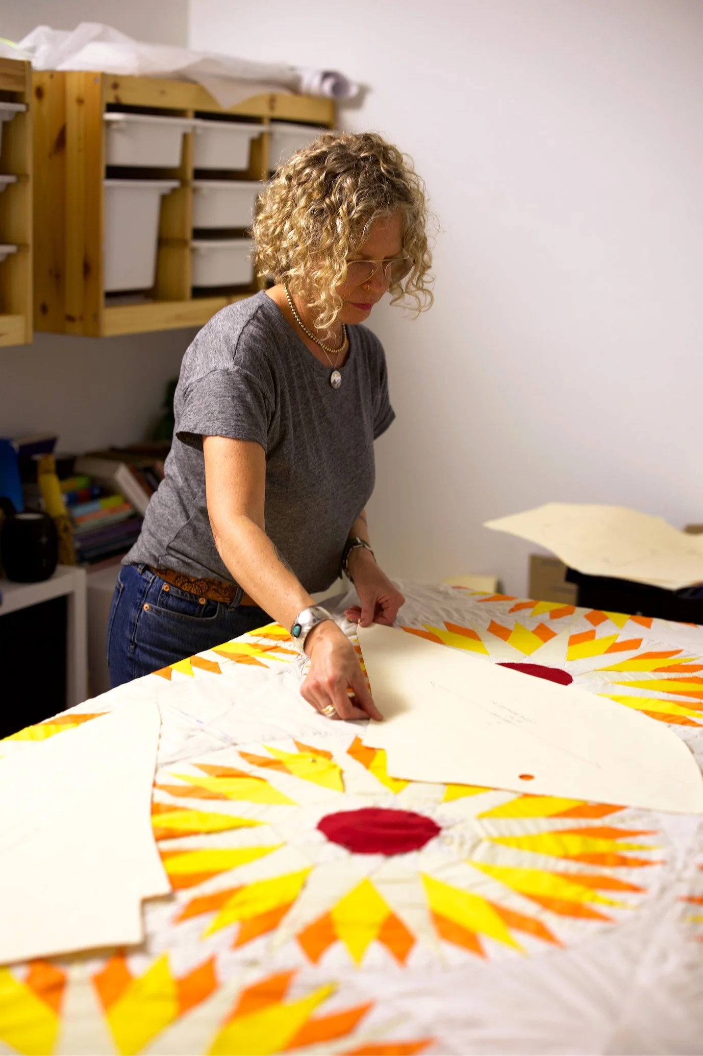 A woman with curly blonde hair, glasses, and wearing a grey shirt and jewelry, is working on a colorful quilt with a sunburst pattern on a table.