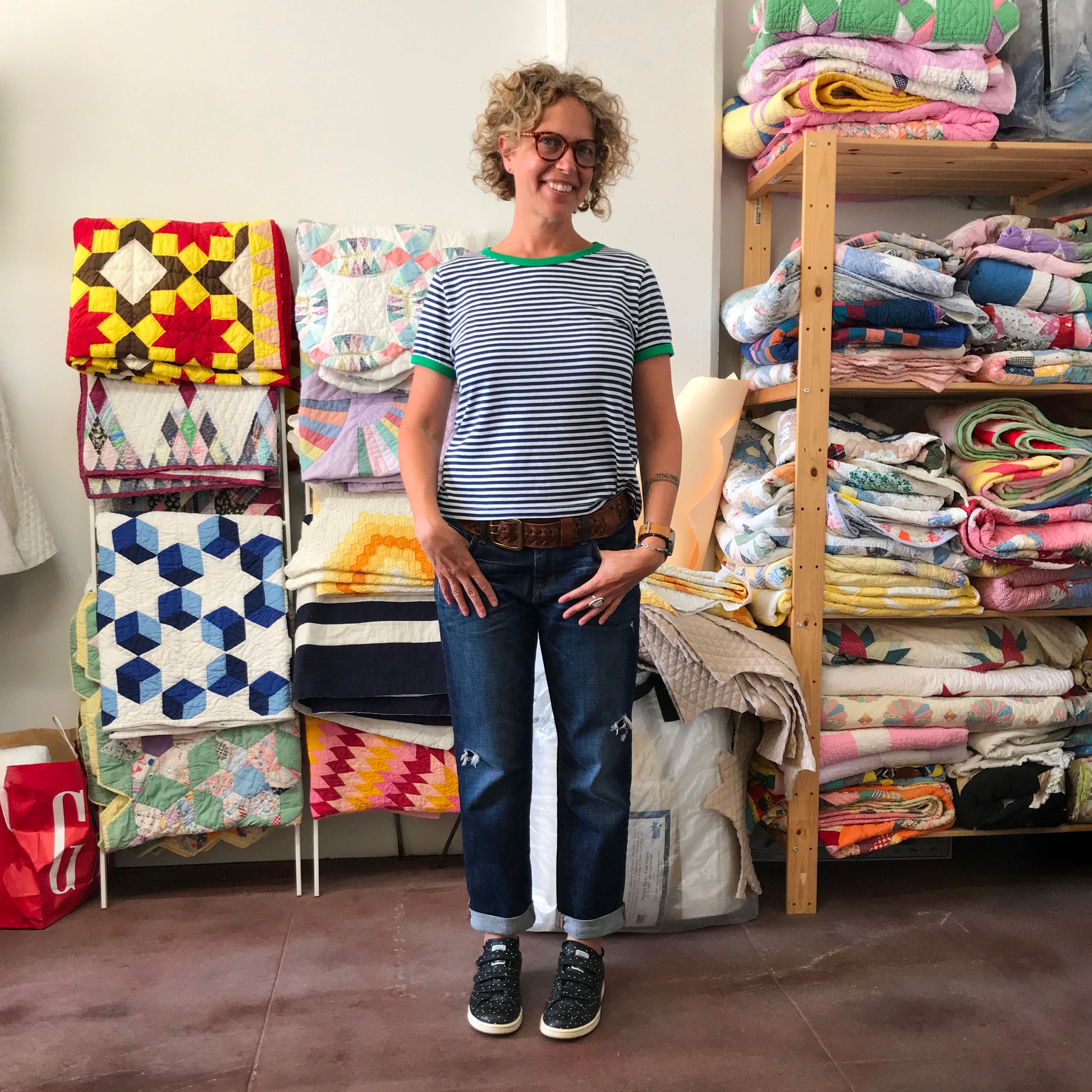Woman with curly blonde hair and glasses standing in front of shelves filled with folded quilts.