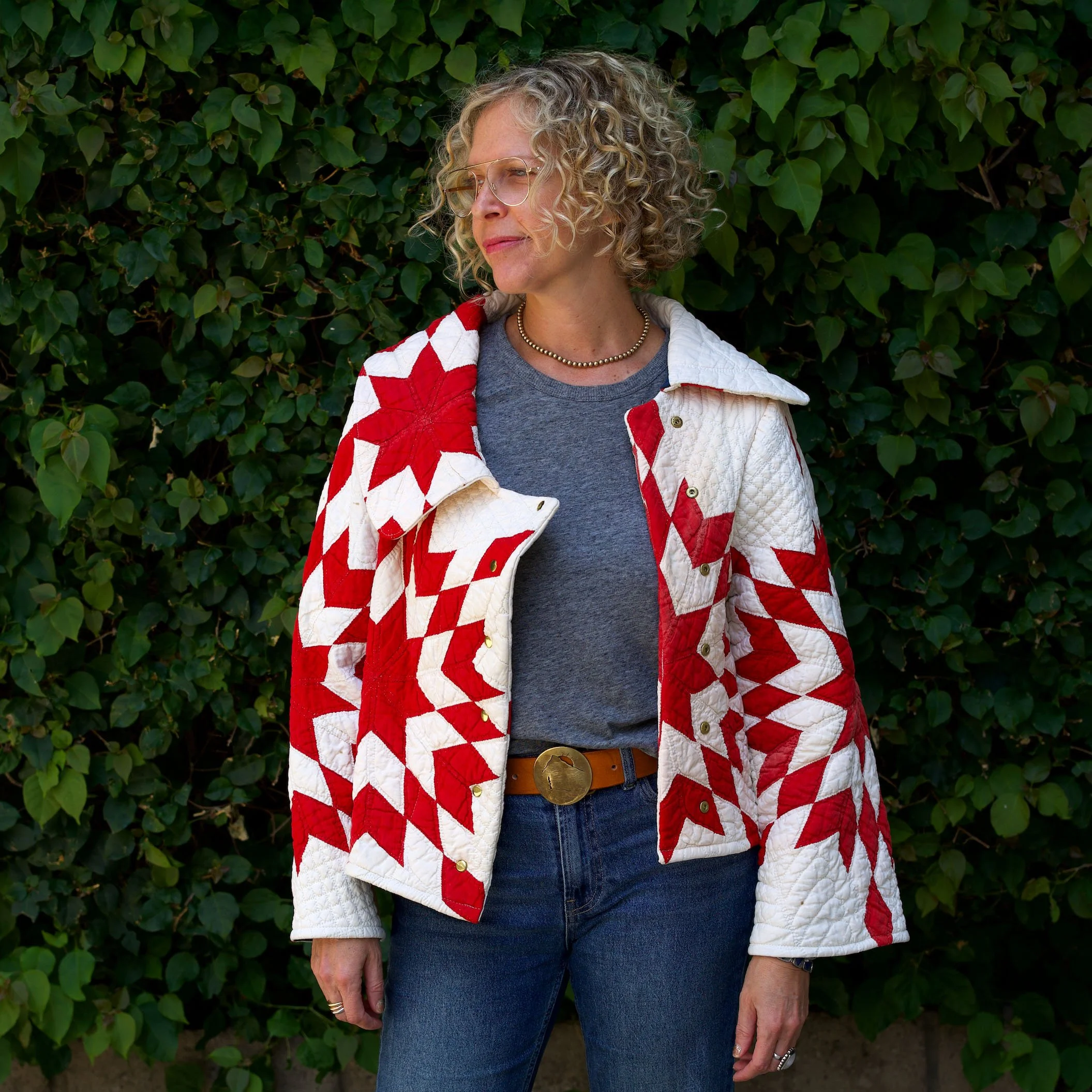 A woman with curly blonde hair and glasses standing outdoors in front of green foliage, wearing a gray shirt, a patriotic red, white, and blue quilted jacket, and blue jeans.